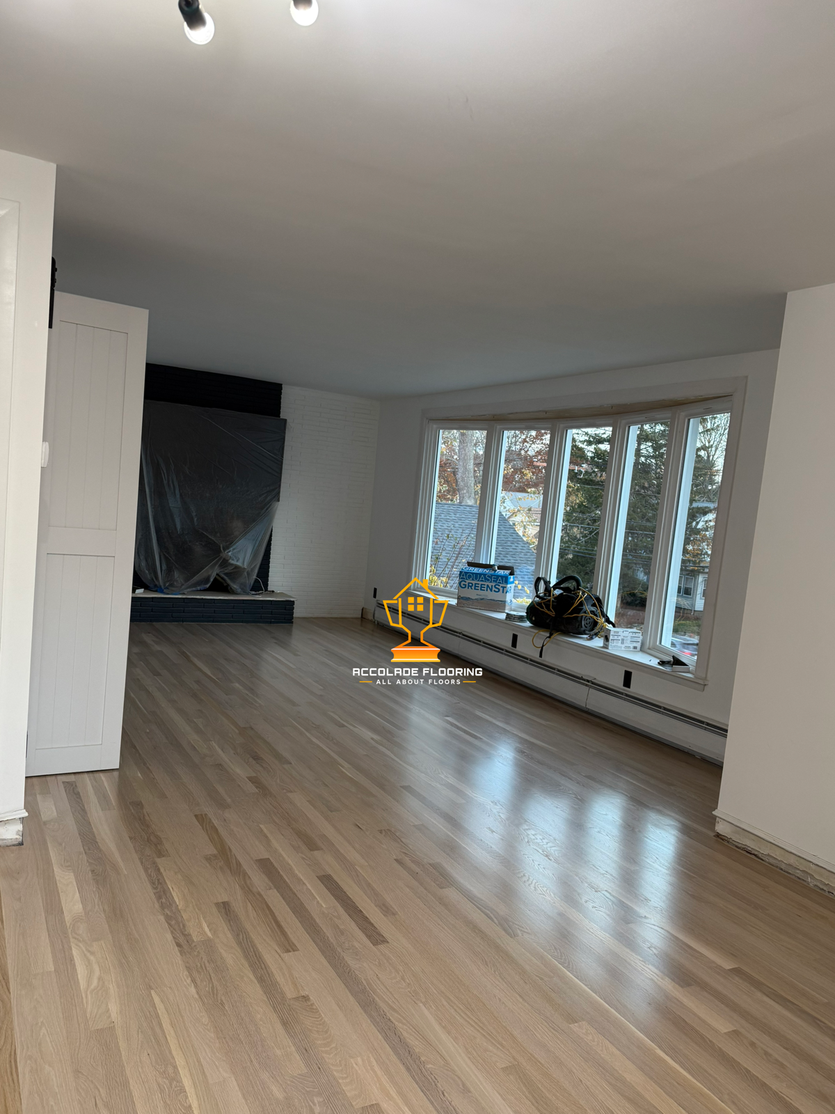 A living room with new light wood flooring, large bay window, and a white brick fireplace covered with a black plastic sheet. Some tools and a bag are placed on the window sill, indicating ongoing renovation.