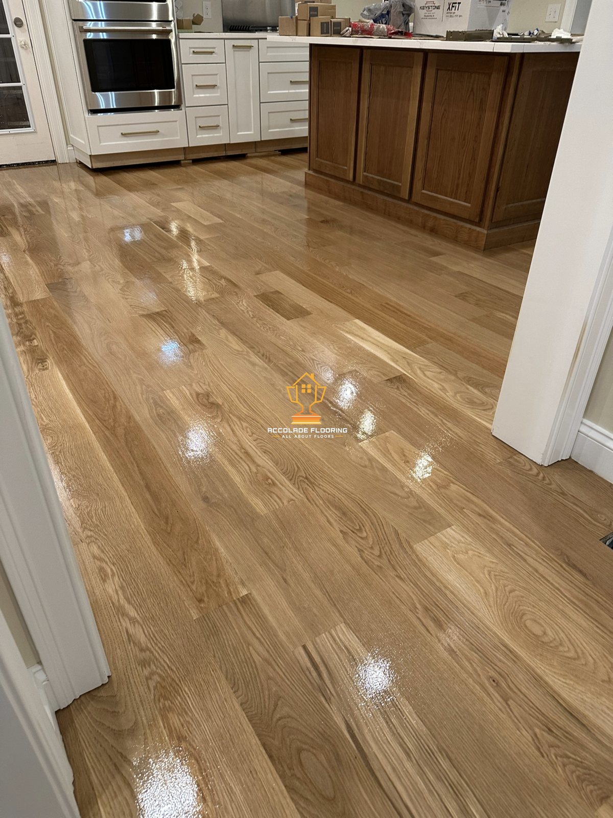 Polished hardwood floor in a kitchen with white cabinets, a stainless steel oven, and a wooden kitchen island with a white countertop. There are boxes and supplies on the countertop. Logo of Accolade Flooring is near the bottom center.