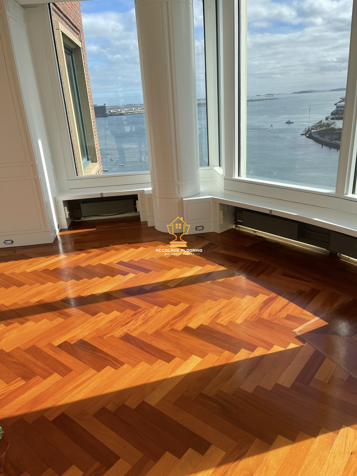 Empty room with large windows overlooking a body of water, with a herringbone patterned hardwood floor and baseboard heating.