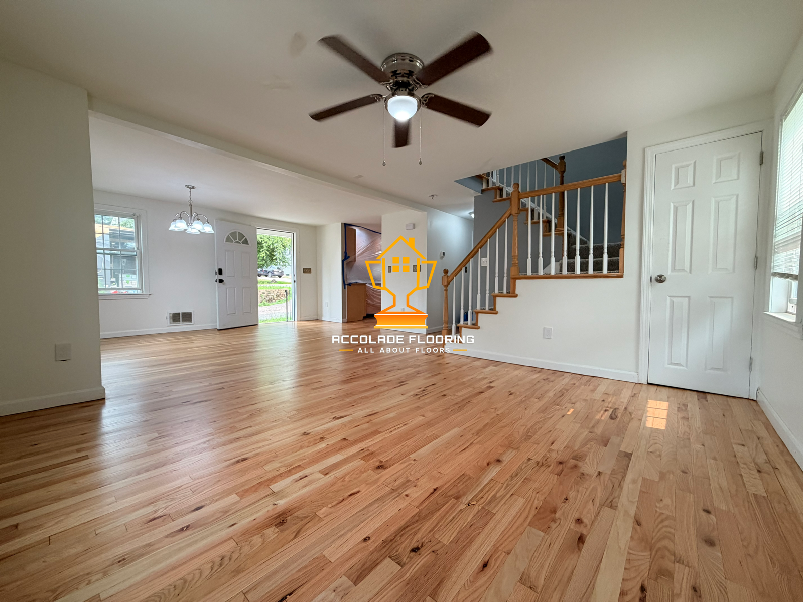Living room with wooden floors, white walls, staircase with wooden banister, window on right, front door, ceiling fan, and chandelier in the background.