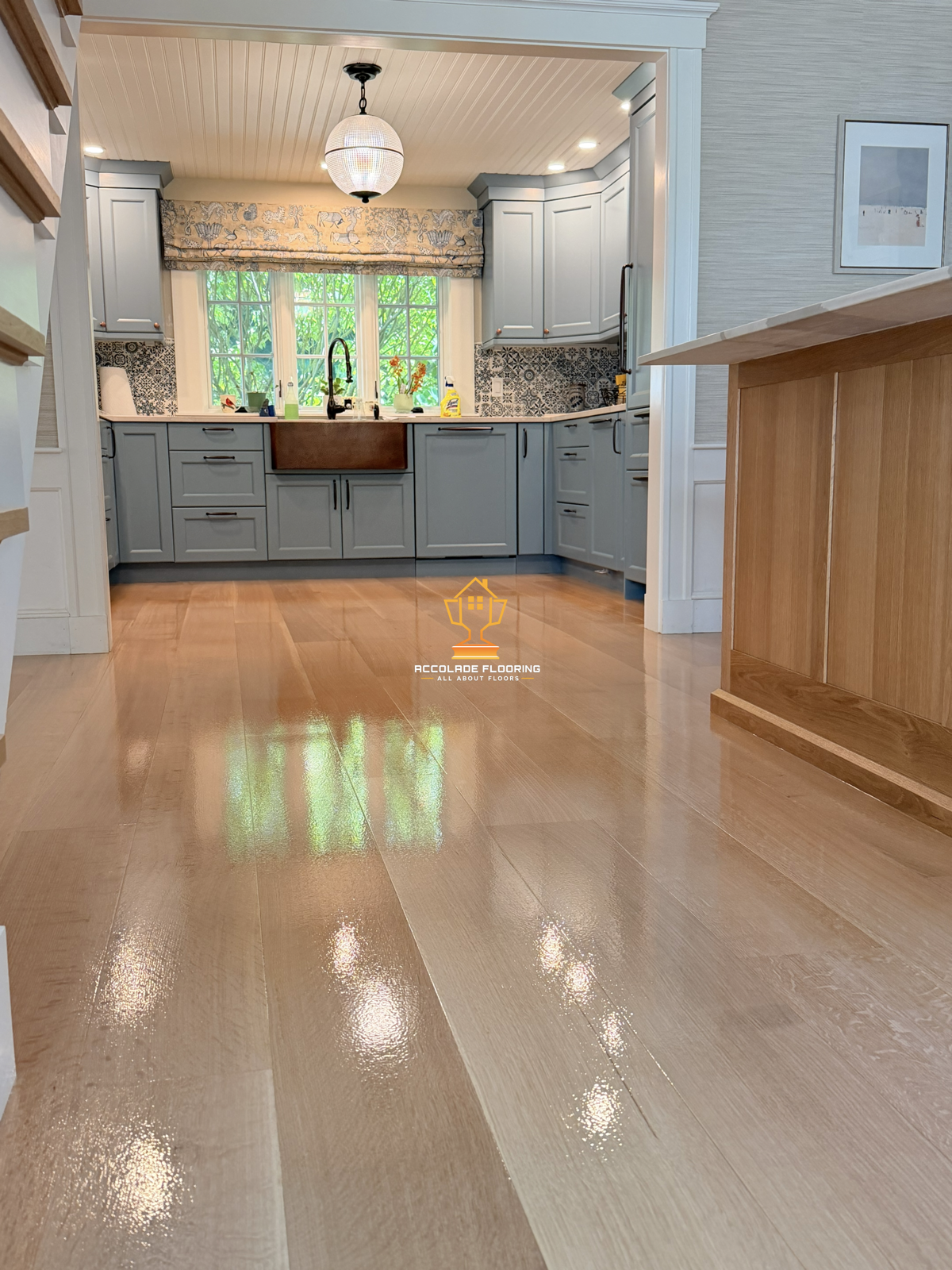 A cozy kitchen with blue cabinets, a large window with greenery outside, a white ceiling with a chandelier, and a wooden floor reflecting light.