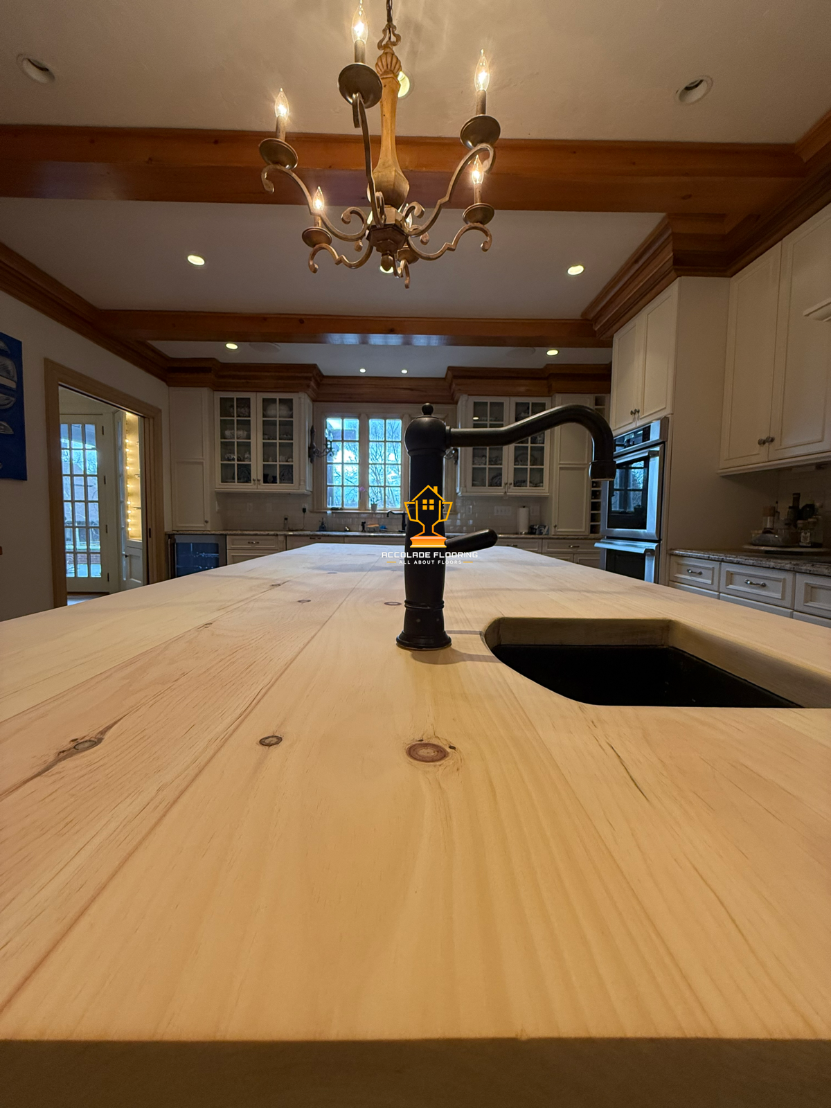 A wooden kitchen island with a black faucet, in a spacious kitchen with white cabinets, a chandelier, and large windows.