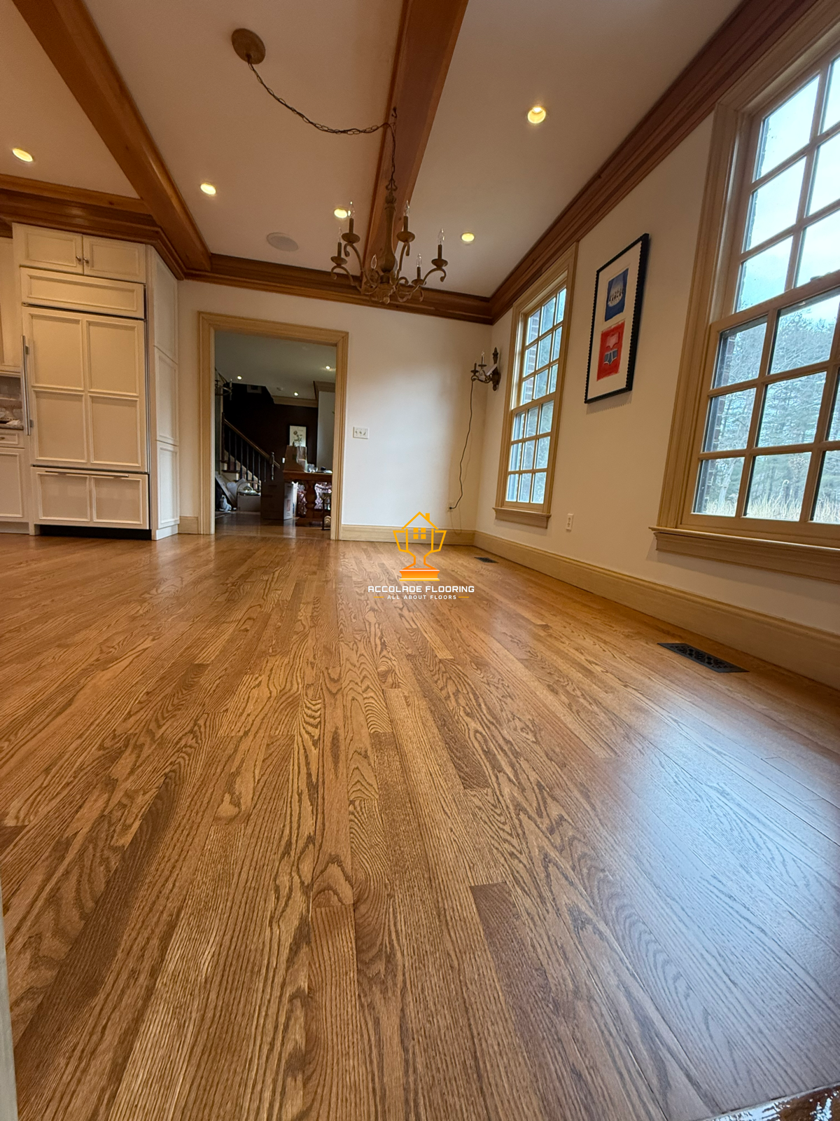 A room with light wooden floors, large windows with wooden trim, a small chandelier, and a doorway leading to another room. The room appears to be empty and newly renovated.