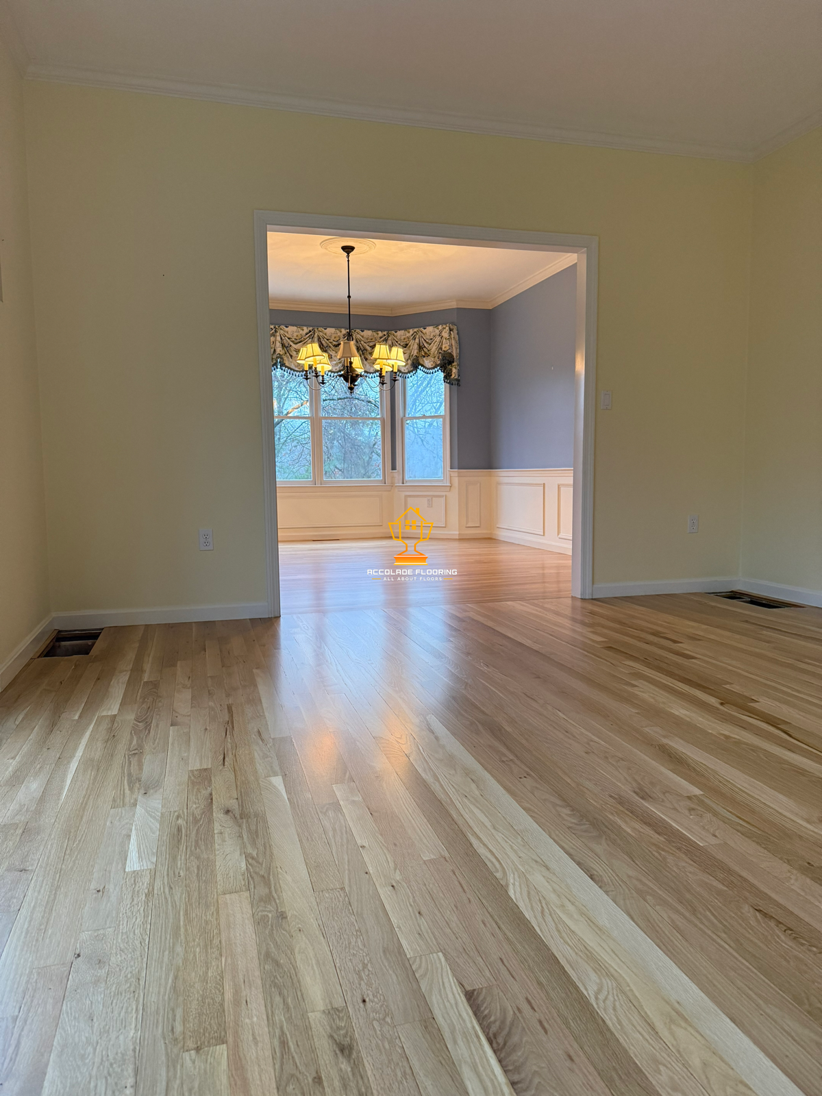 Empty living room with hardwood floors, light yellow walls, and a doorway leading to a dining area with a chandelier and large windows.