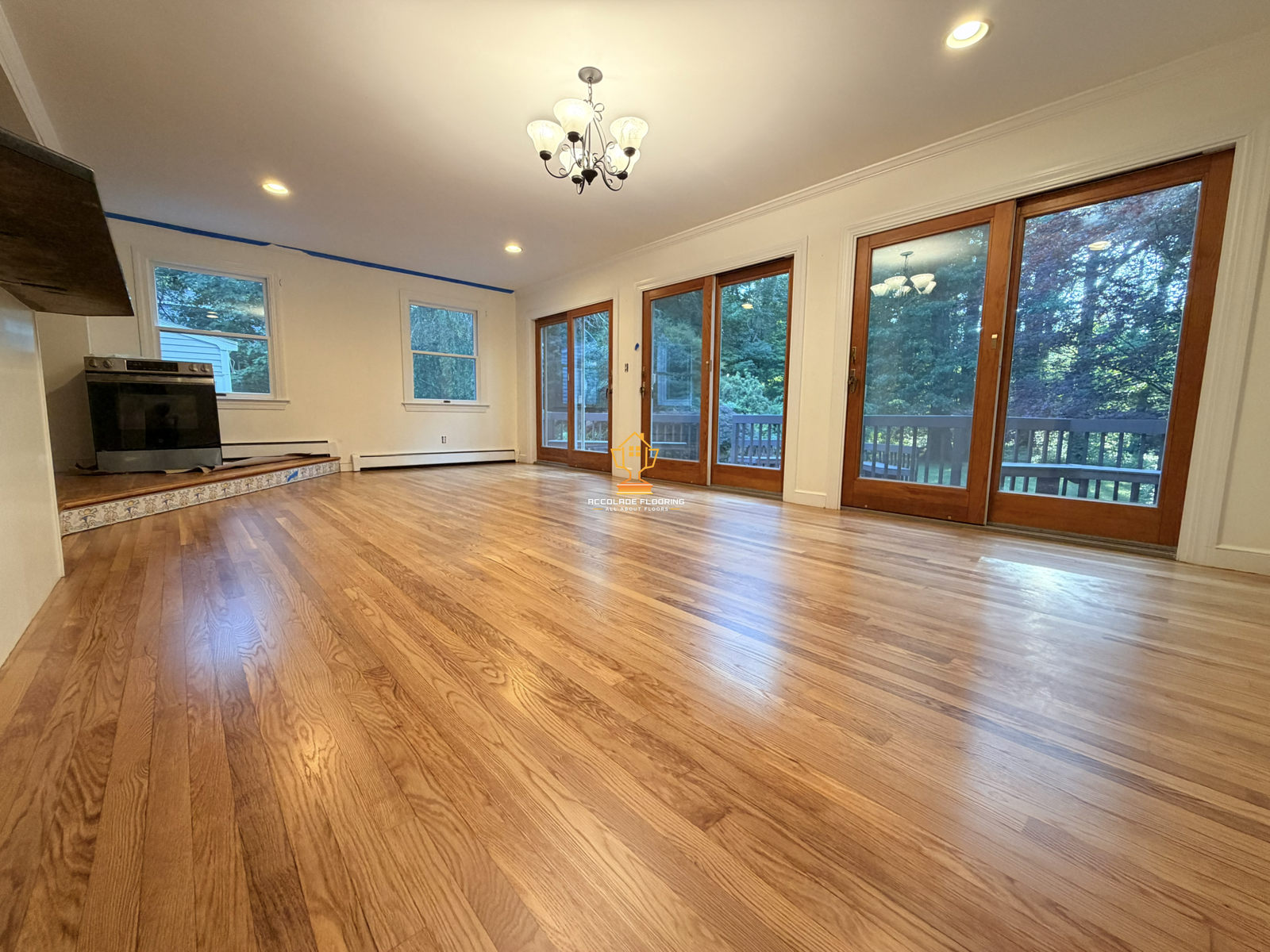 Empty living room with hardwood floors, large windows, sliding glass doors leading to a deck, and a chandelier.