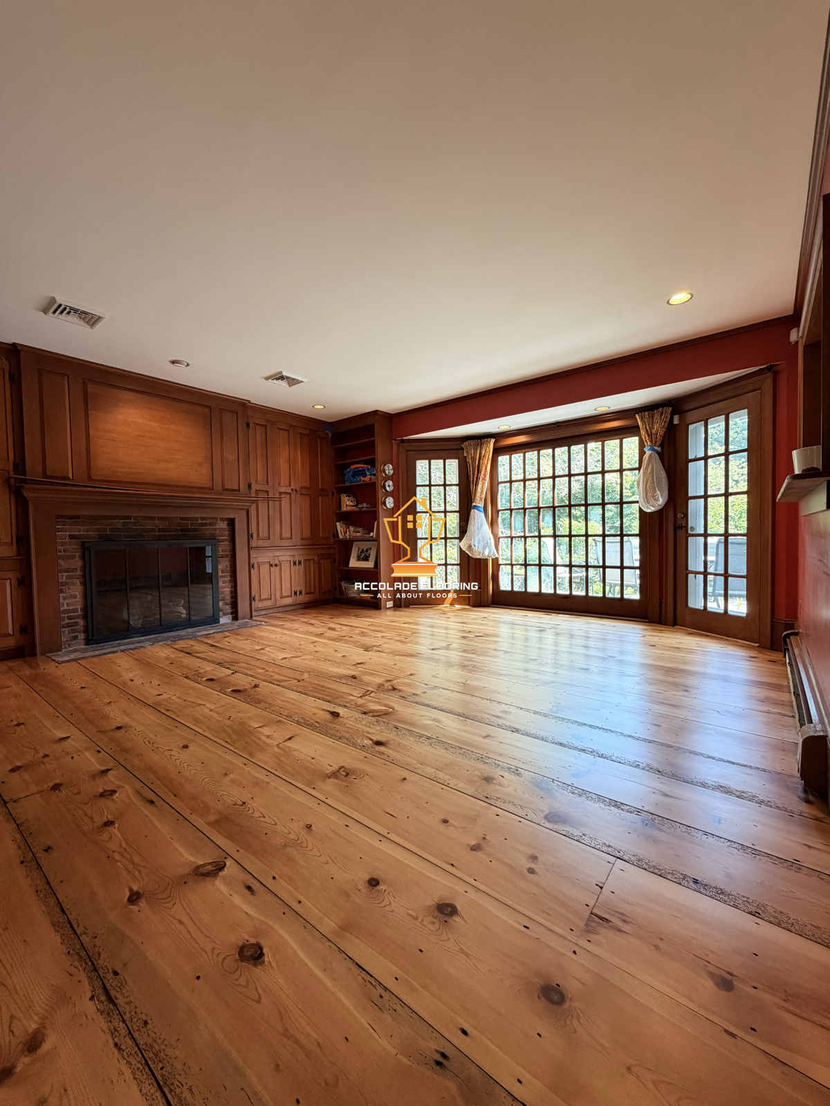 Empty living room with wooden floors, wood-paneled walls, a brick fireplace, a built-in bookshelf, and large glass doors with curtains leading to the outside.