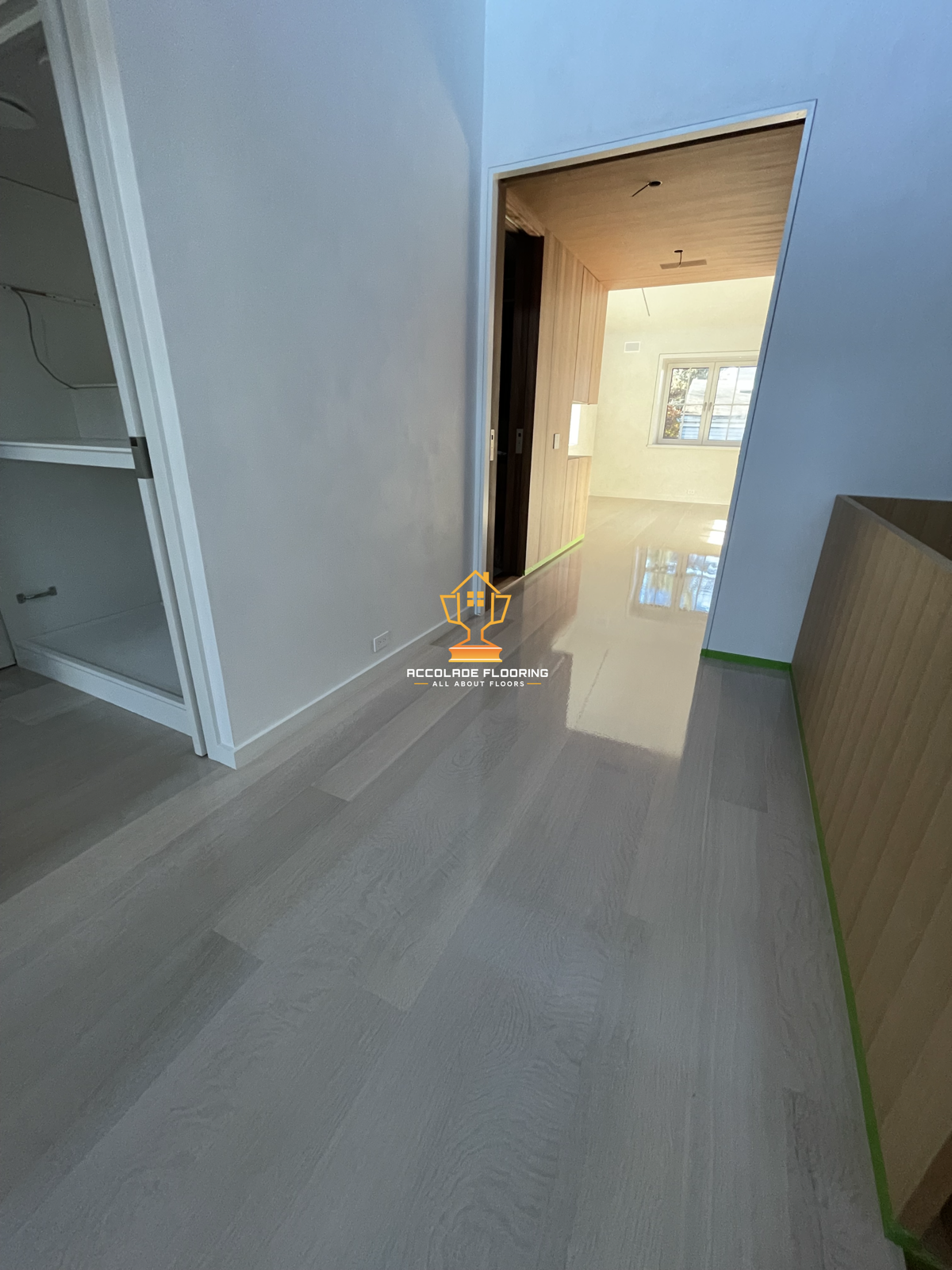 Interior view of a room with light wood flooring, white walls, and a partial view of a kitchen or hallway with wood paneling, and a window allowing natural light inside.