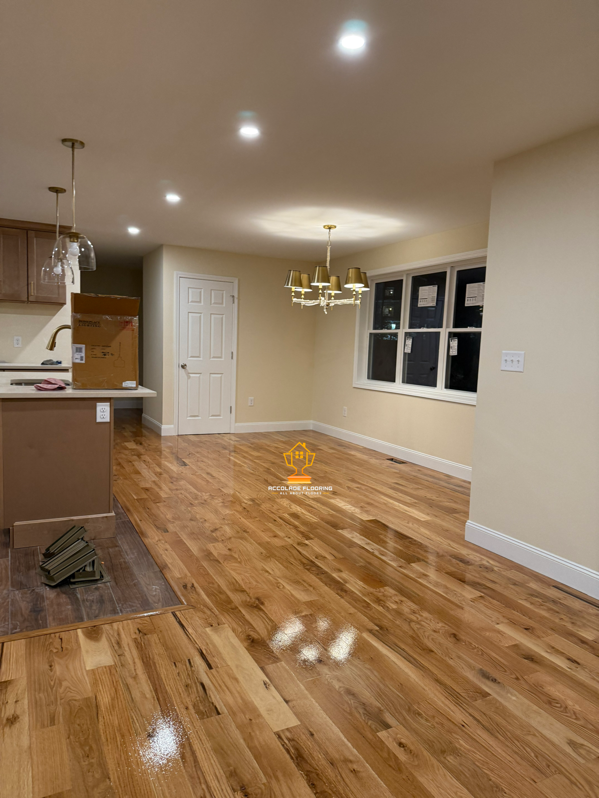 Empty room with newly installed hardwood floors, chandelier, large window, and a kitchen area with pendant lights.