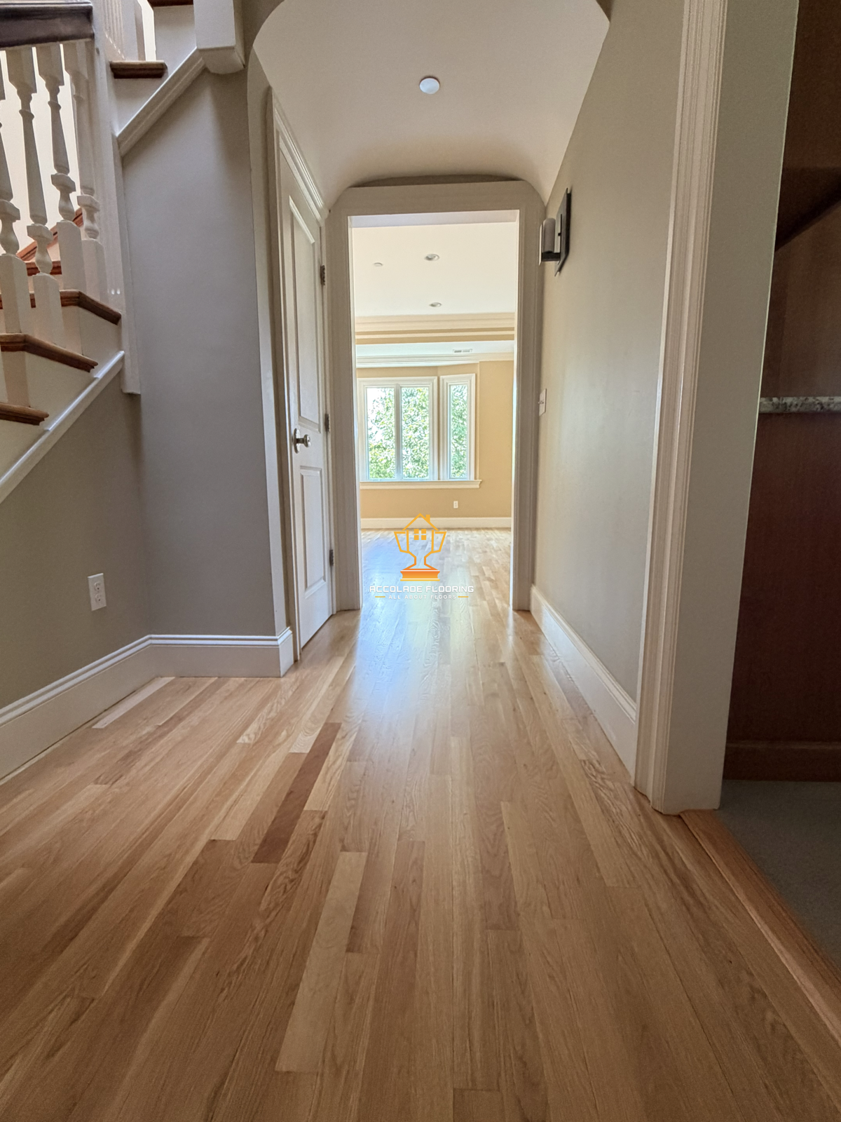 A hallway with light-colored wooden flooring and cream-colored walls leads to a room with a large window letting in natural light.