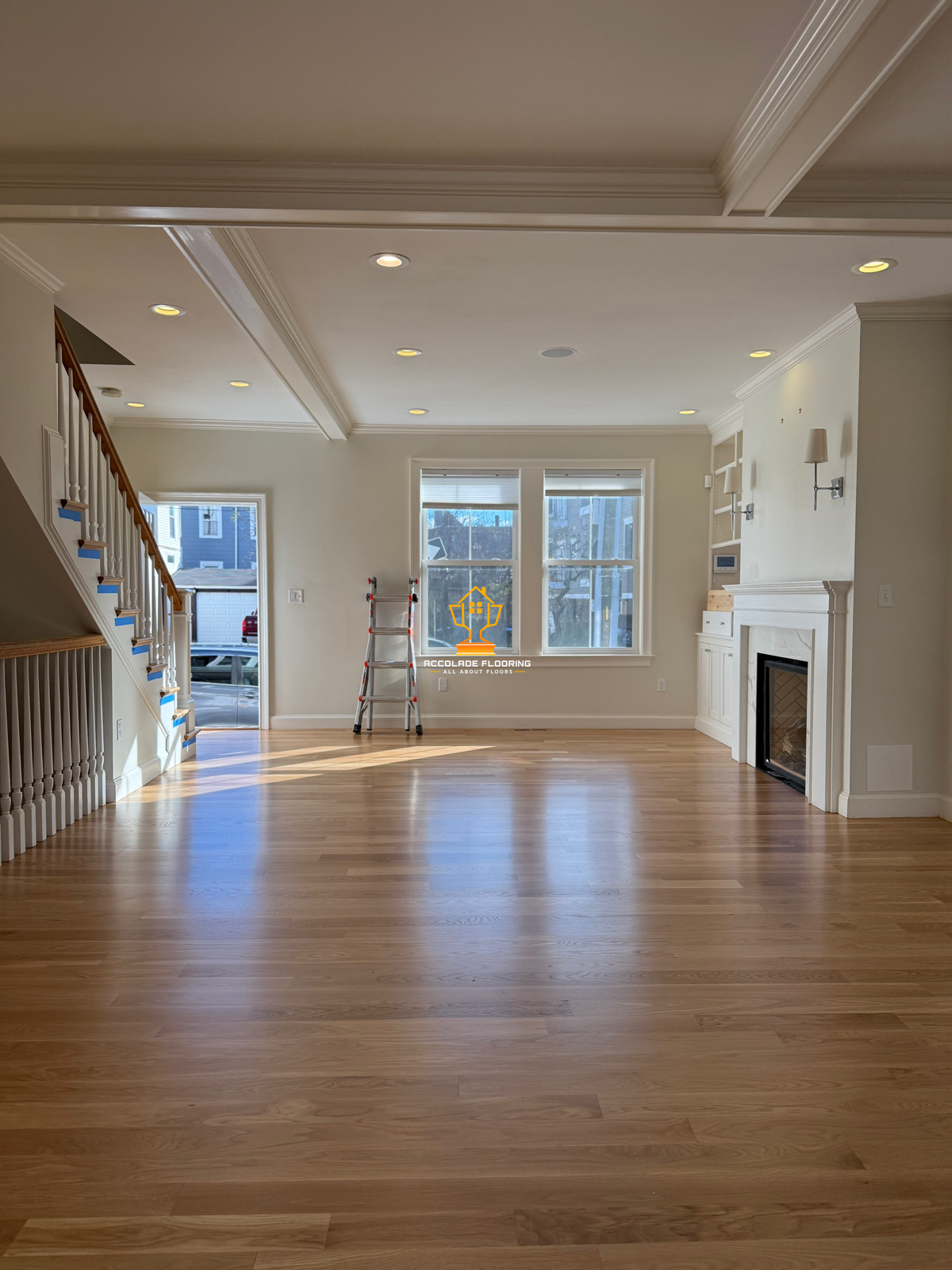 An empty living room with light hardwood floor, white walls, and a fireplace. There are three large windows and a doorway leading outside. A ladder is near the window, and the ceiling has recessed lighting.