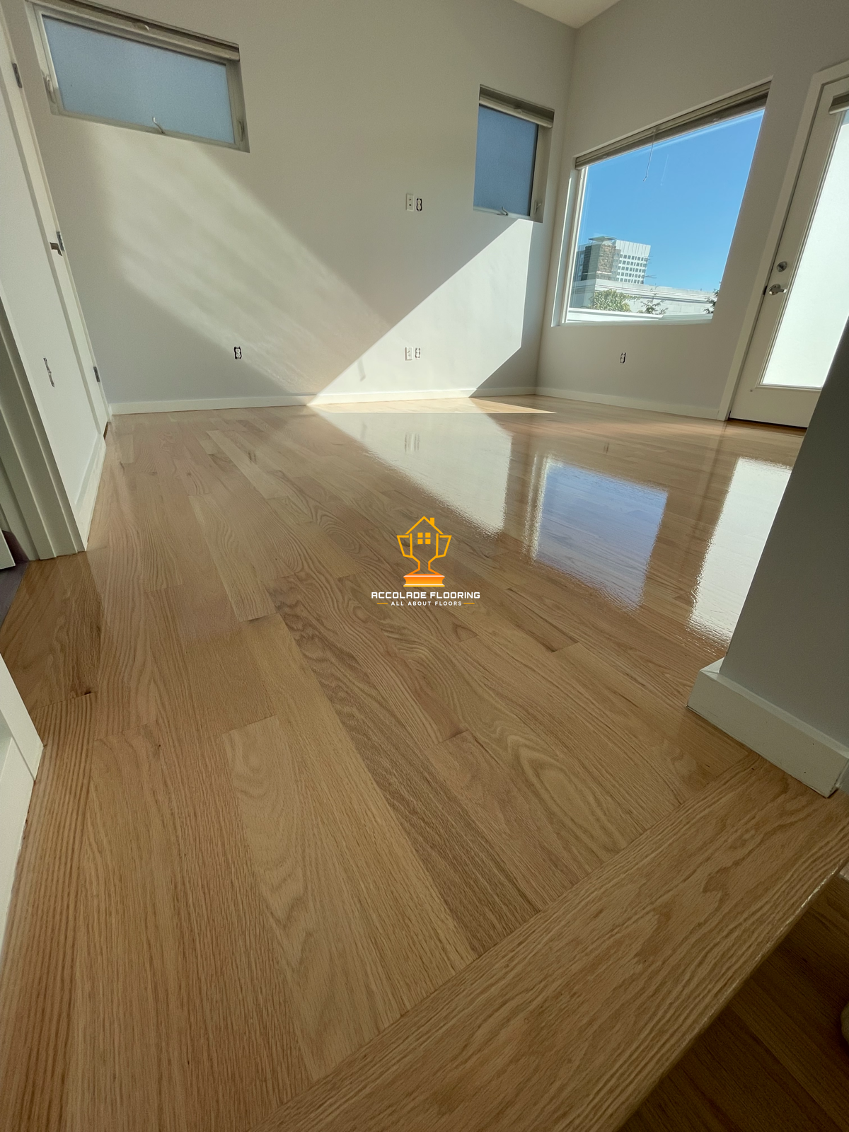 Empty room with large windows letting in sunlight, showcasing newly installed, polished oak flooring with a natural wood grain pattern.