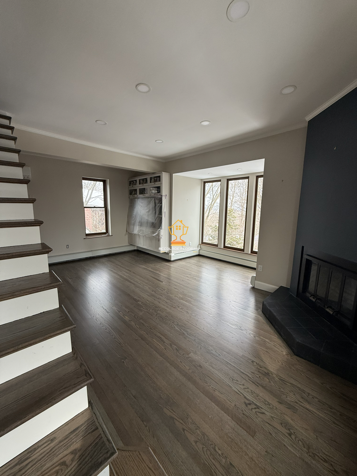 Empty living room with hardwood floors, large windows, a blue accent wall, and a black fireplace.