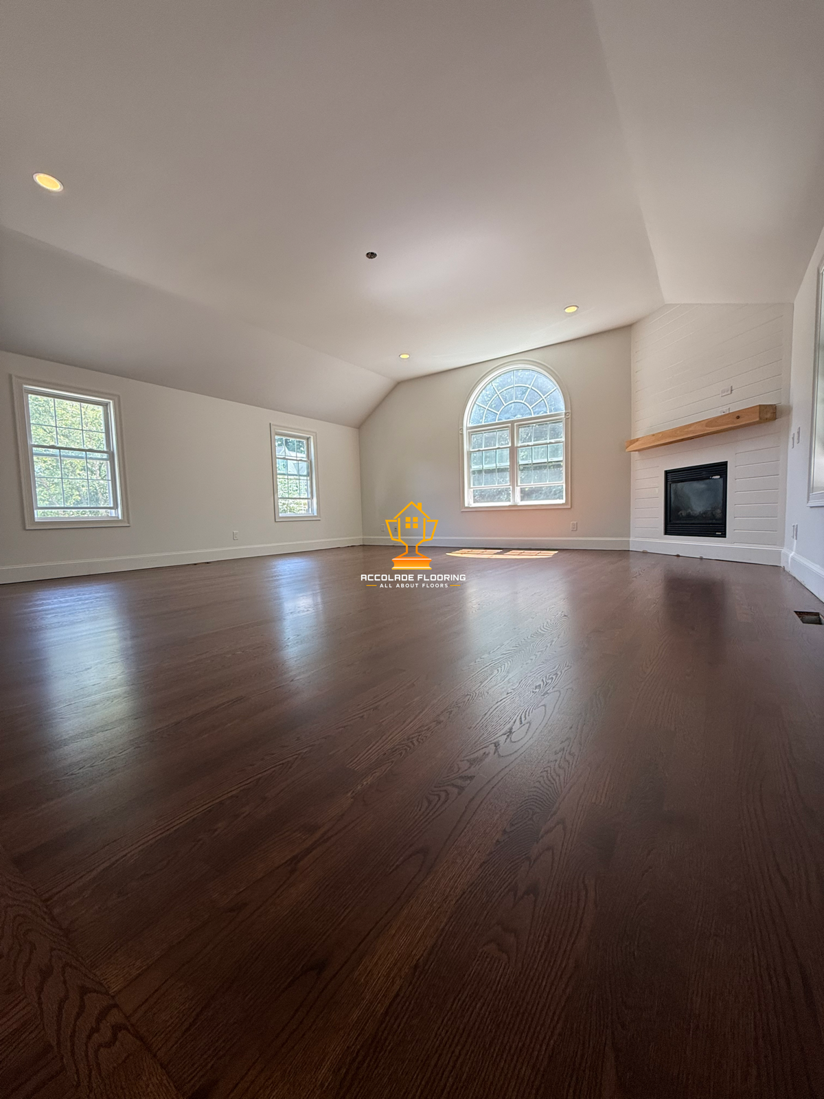Empty living room with hardwood floors, white walls, three windows, a fireplace with a wooden mantel, and an arched window.