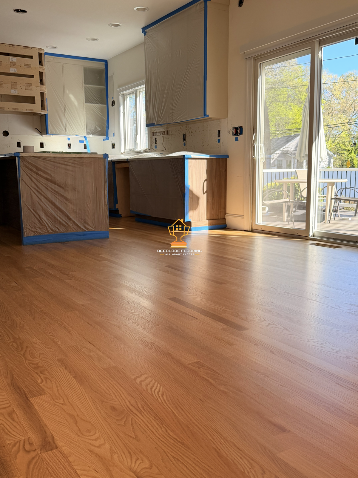 Room with newly installed hardwood flooring, partially covered cabinets, and a sliding glass door leading to an outdoor balcony with patio furniture. Windows let in natural light.