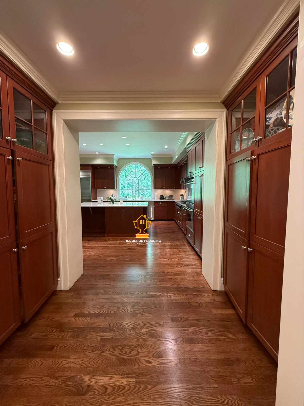 View through a doorway into a kitchen with dark wood cabinets, hardwood floors, and a large window at the sink area.