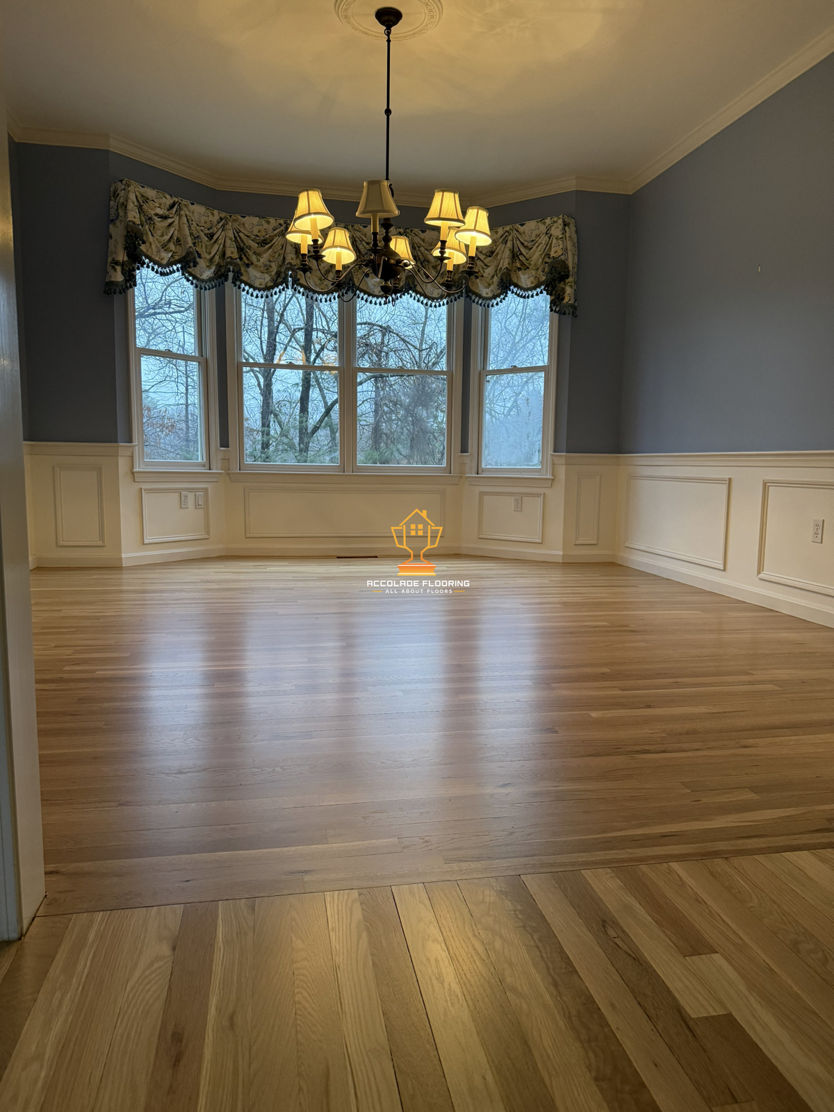 Empty living room with hardwood flooring, blue walls, and large windows with floral valances. Chandelier hanging from ceiling.