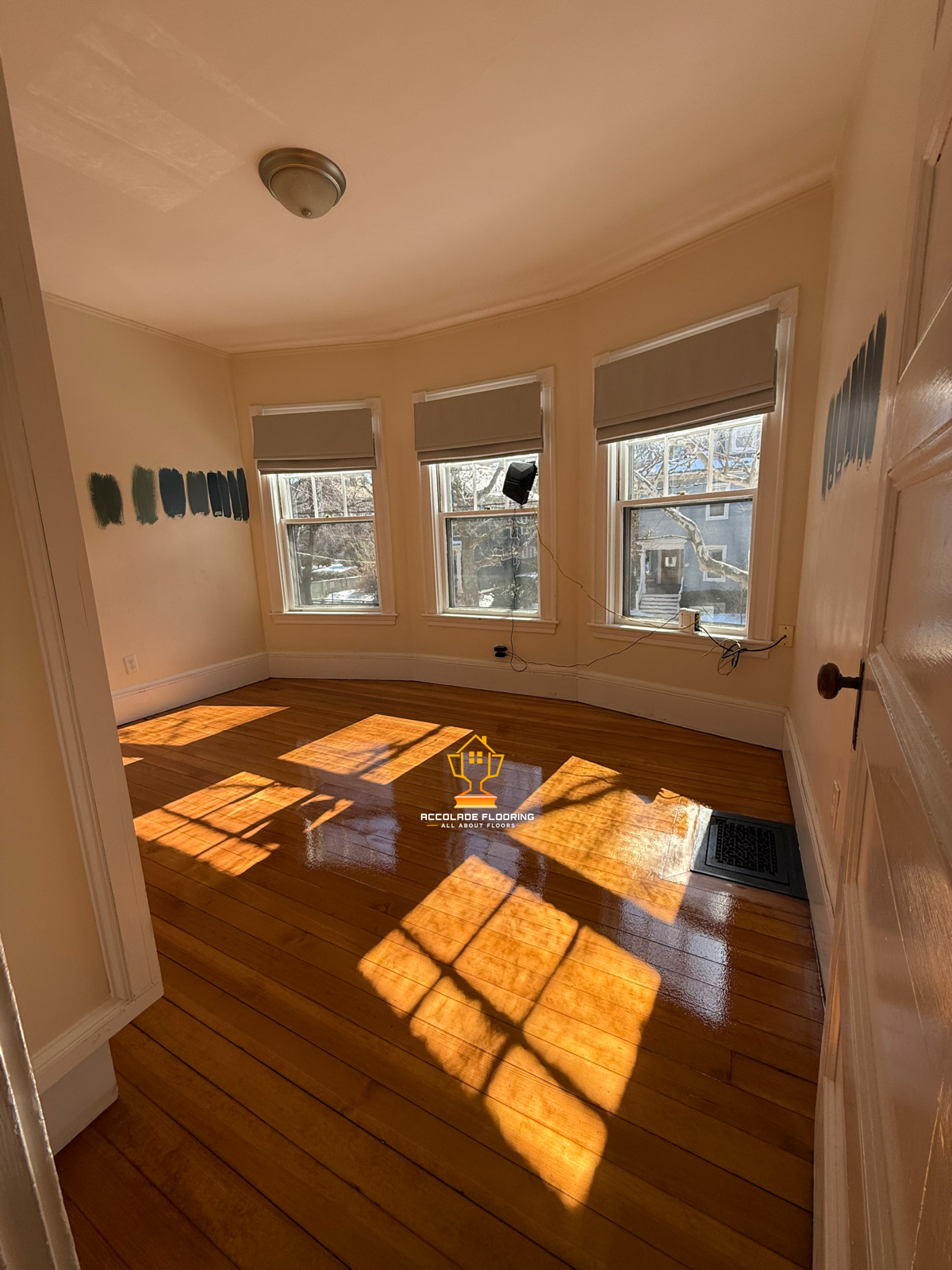 Empty room with three windows allowing sunlight in, hardwood floor with sunlight shadows, beige walls, and paint swatches on one wall.