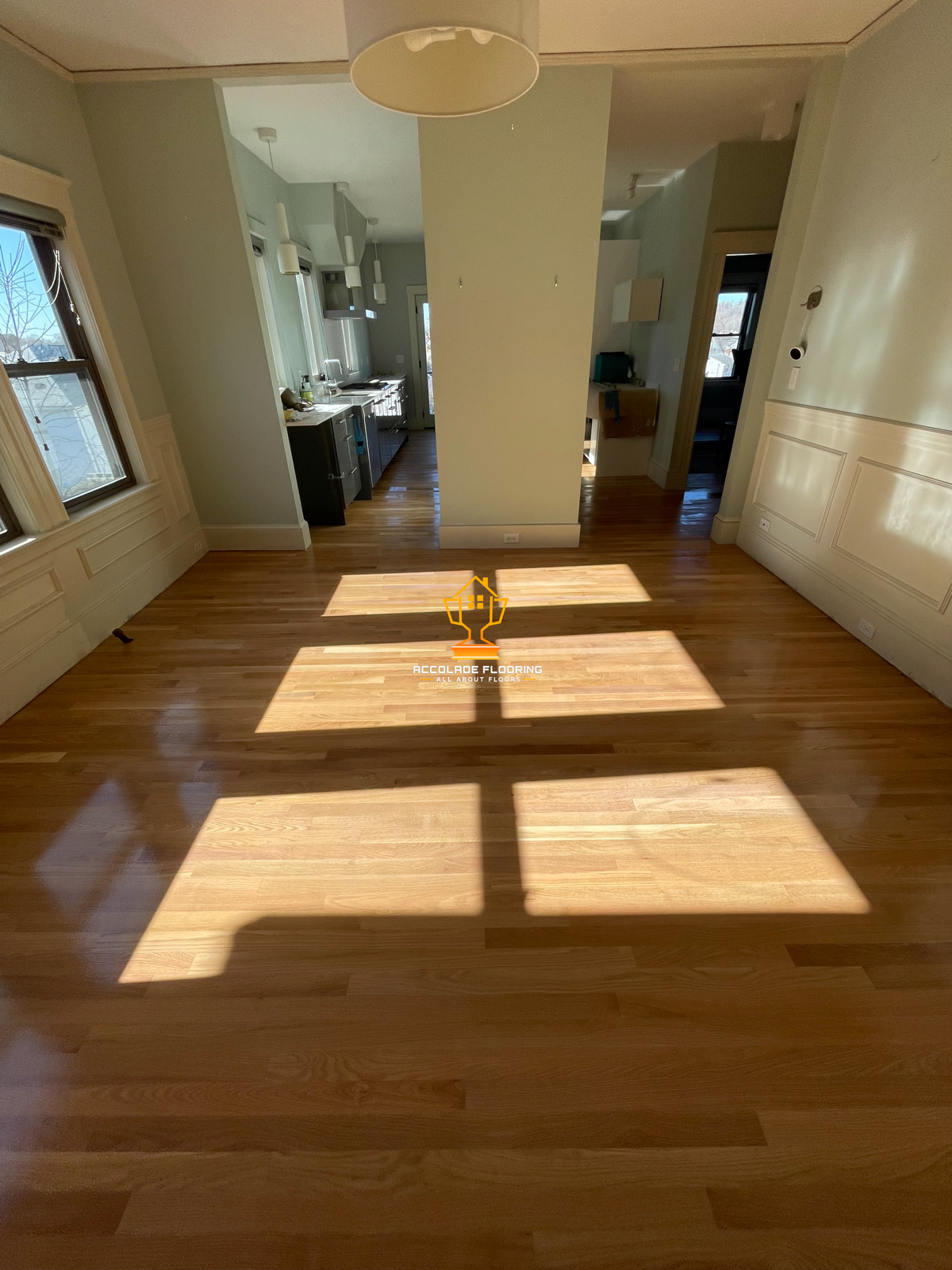 Empty living room with hardwood floors and sunlight coming through windows, casting shadows on the floor, with kitchen visible in the background.