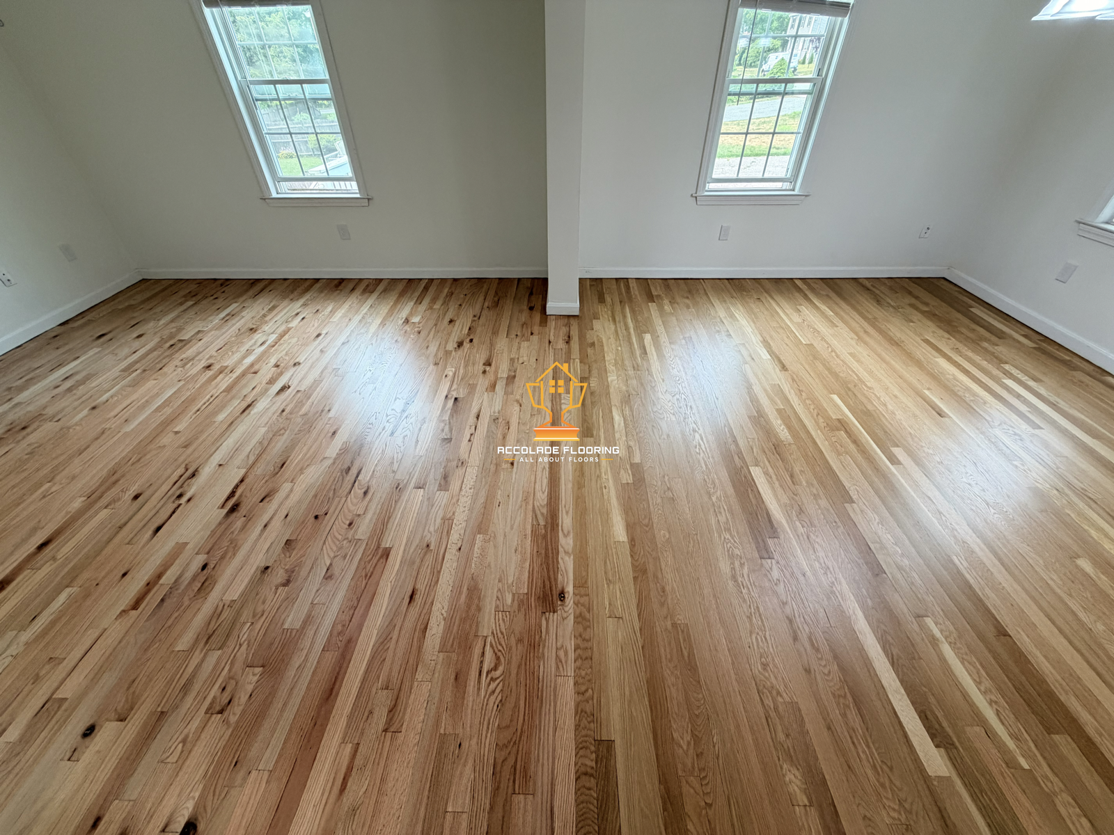 Empty room with newly installed light wood flooring, white walls, and three large windows allowing natural light, with a central pillar dividing the room.
