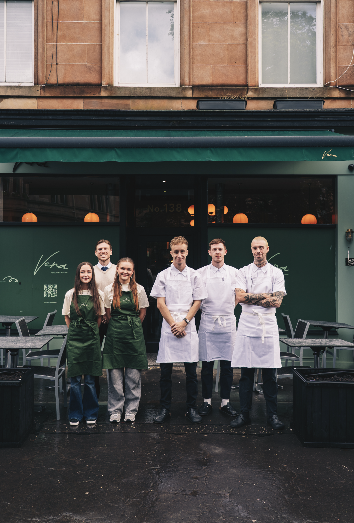 Group of six restaurant staff members standing outside the green restaurant with the name 'Vera'. Four are in white chef uniforms, two are in green aprons and casual clothing, smiling in front of the restaurant entrance. Served at Vera Restaurant Gla