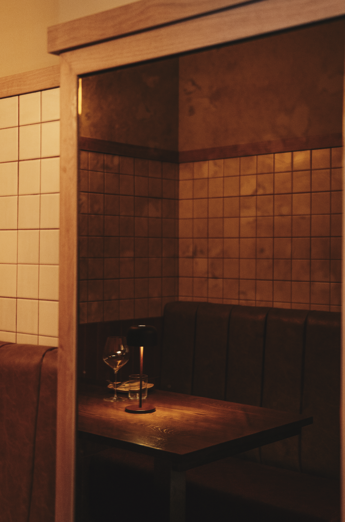 A cozy restaurant dining area with a wooden table, wine glass, and black lamp, framed by tiled walls and padded booth seating. Served at Vera Restaurant Glasgow Southside 138 Nithsdale Road.