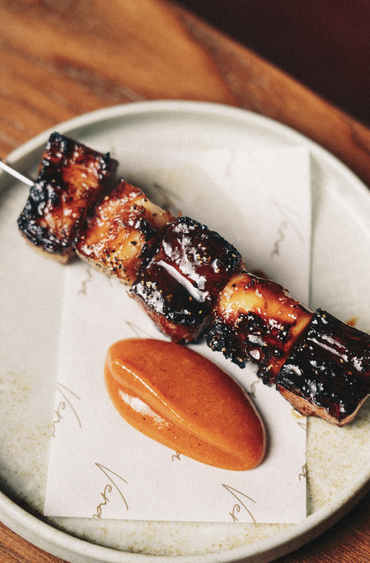 Barbecue ribs on a skewer served alongside a tomato slice on a white plate with decorative writing, placed on a wooden table. Served at Vera Restaurant Glasgow Southside 138 Nithsdale Road.