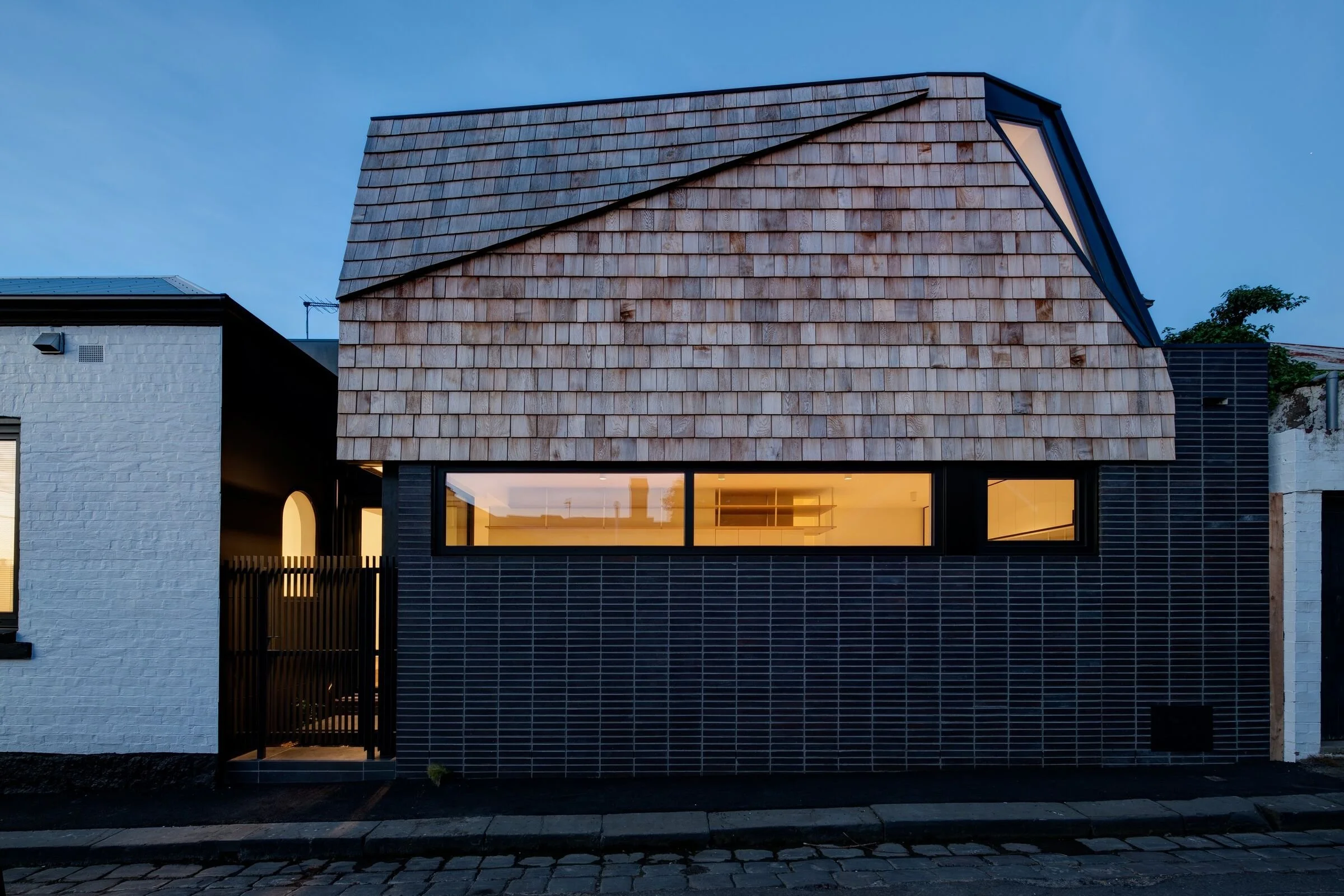Modern house with a unique sloped wooden shingle roof, black brick facade, large window, and a small side gate, lit from inside during twilight.