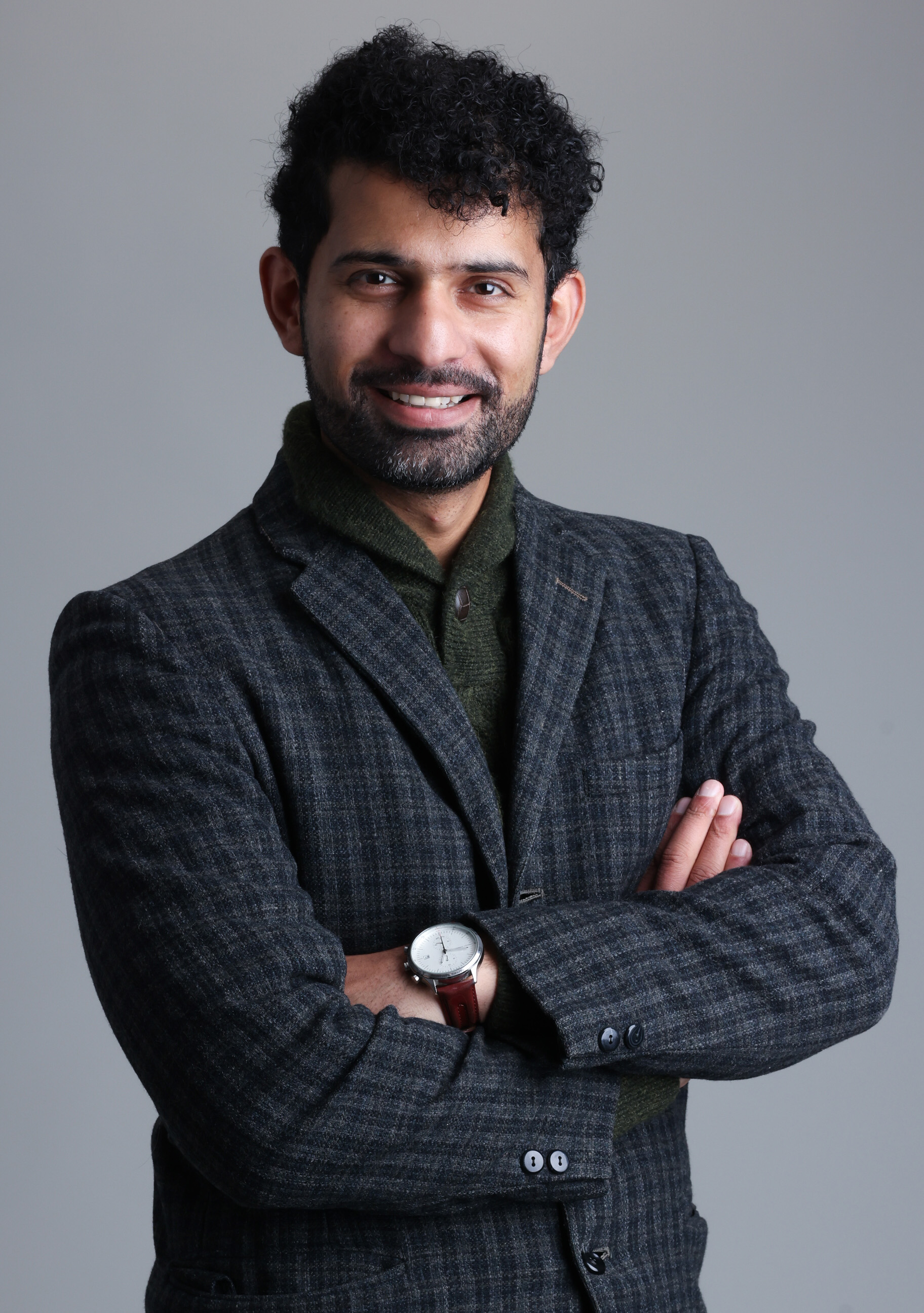 A man with dark, curly hair and a beard, wearing a dark plaid blazer over a green sweater, standing with arms crossed and smiling at the camera against a plain gray background.