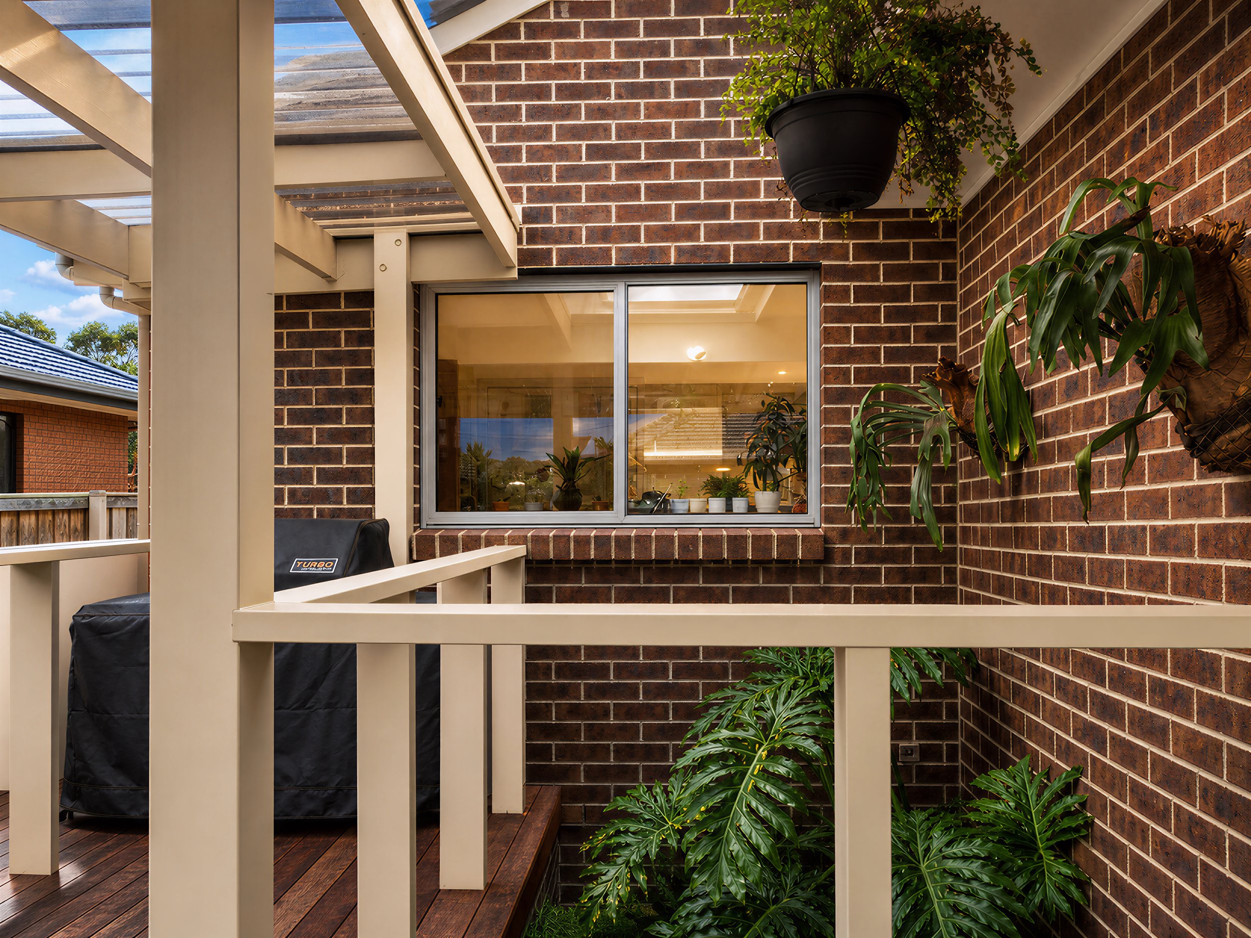 View of a balcony with a beige railing, a barbecue cover, and indoor plants near a brick wall with a window reflecting the sky and clouds.