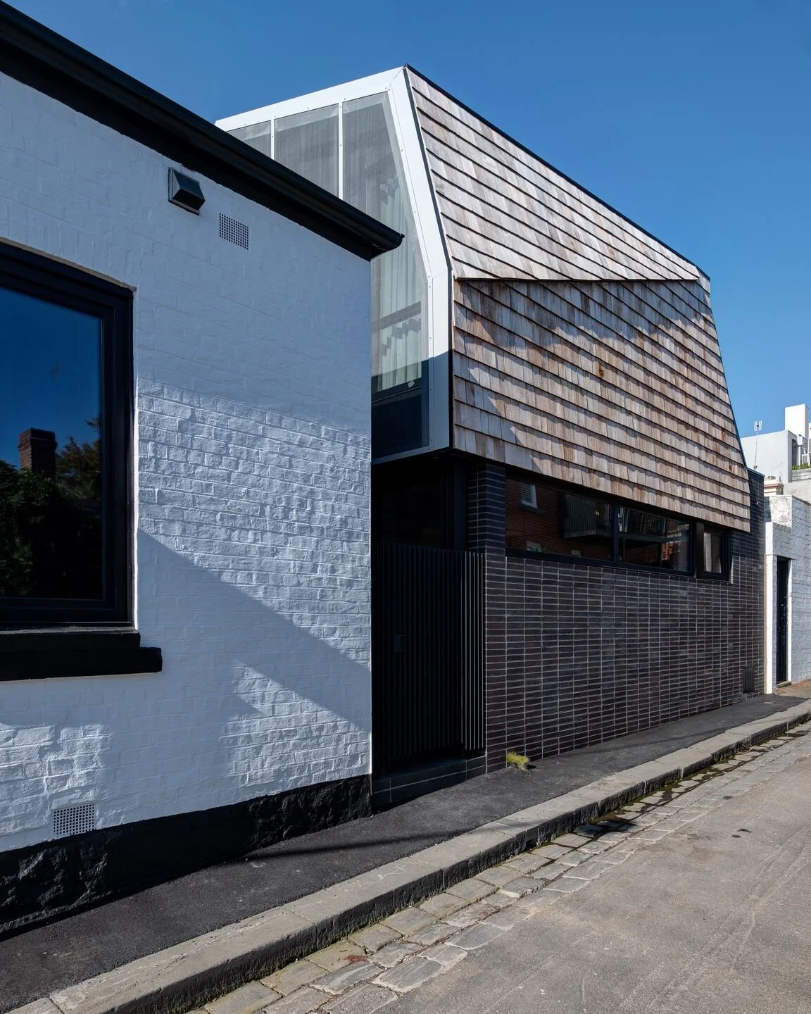 Modern building with white painted brick wall, black window frame, and upper section covered with weathered wooden shingles, under a clear blue sky.