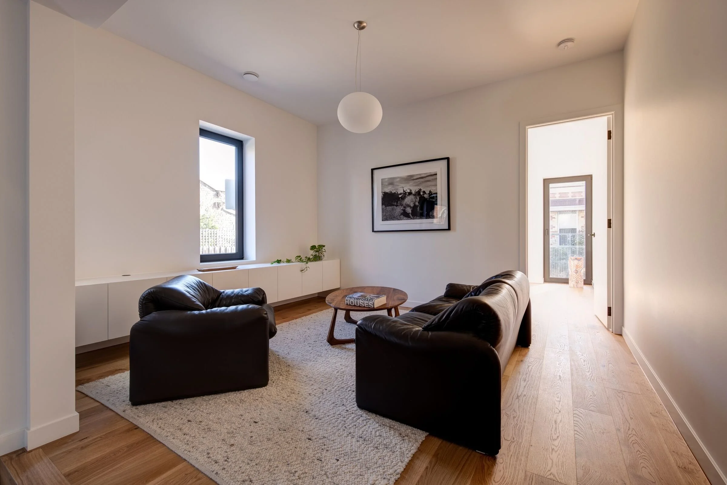 Living room with three black leather sofas, a round wooden coffee table holding a book, a cream-colored rug, a white built-in cabinet with a small green plant, a black and white framed photograph of a dog, a window, a door leading outside, a pendant light, and light wood flooring.
