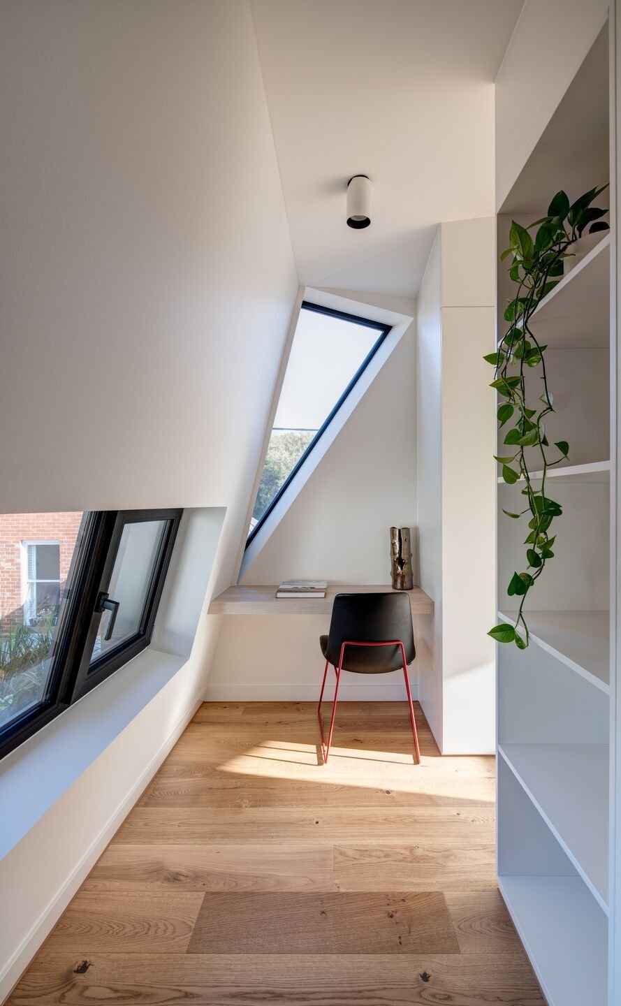 A minimalist home office with a slanted ceiling, a rectangular window, a small wooden desk with books and a decorative vase, a black chair with red legs, a white bookshelf with a hanging plant, and wooden flooring.