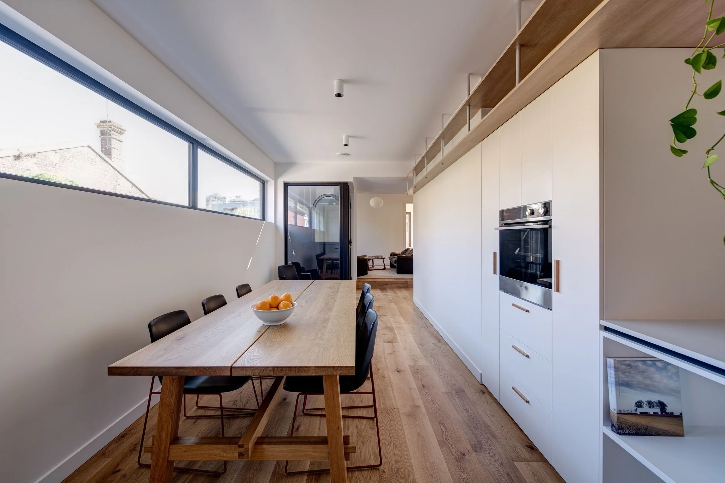 Modern dining area with wooden table, black chairs, white cabinetry, built-in oven, large horizontal window, and wooden flooring in a minimalistic kitchen.