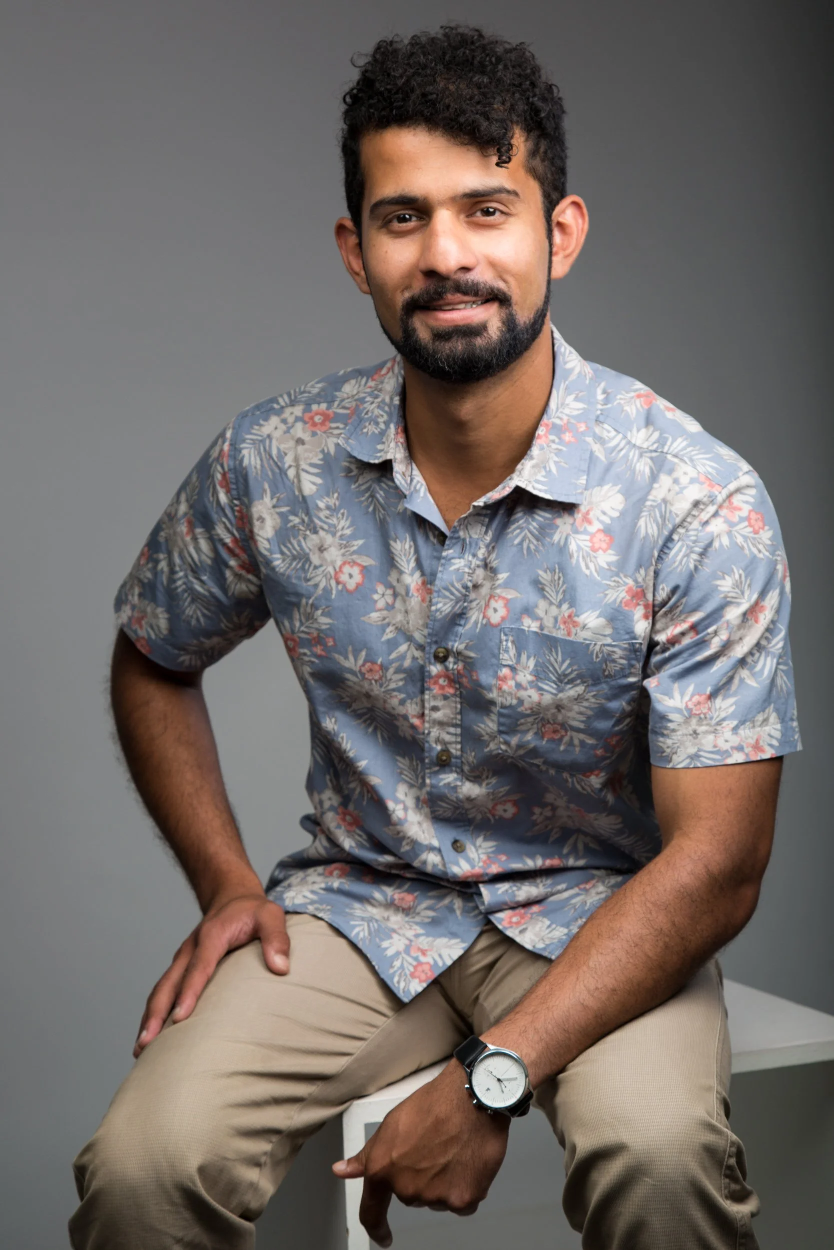 A man with curly hair and a beard wearing a floral shirt and beige pants, sitting on a white stool against a gray background, looking at the camera and smiling