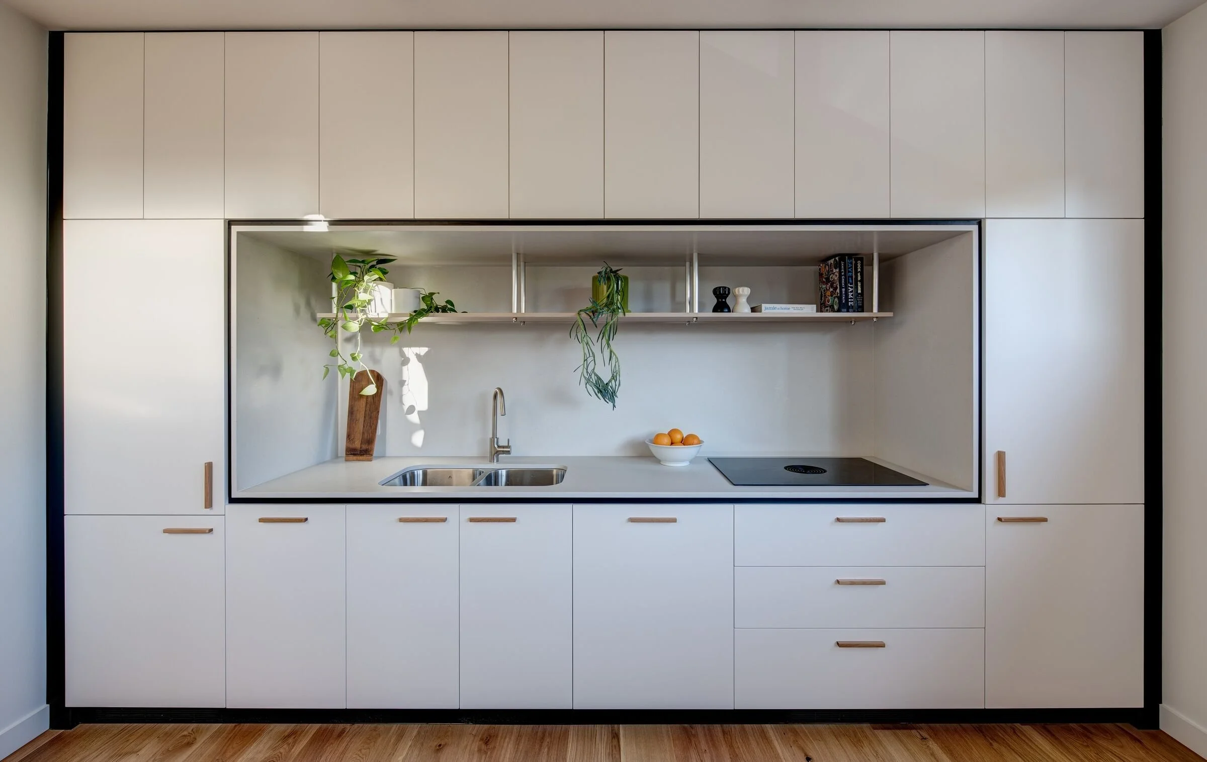 Modern minimalist kitchen with white cabinetry, open shelving, a bowl of oranges, potted plants, and a stovetop.