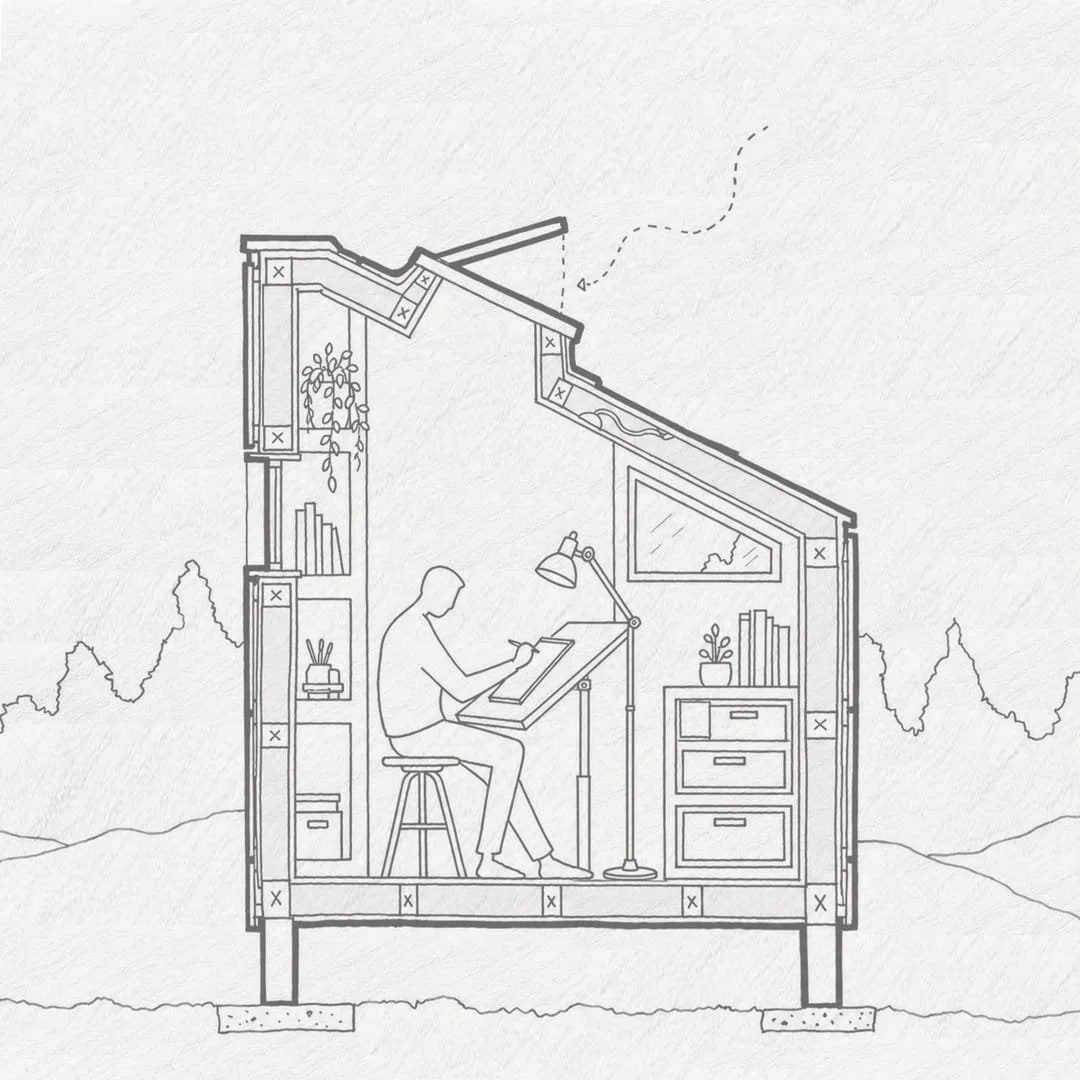 Cross-section of a small house with a person sitting at a drafting table under a sloped roof, bookshelves, a desk with drawers, and a lamp, with trees outside.