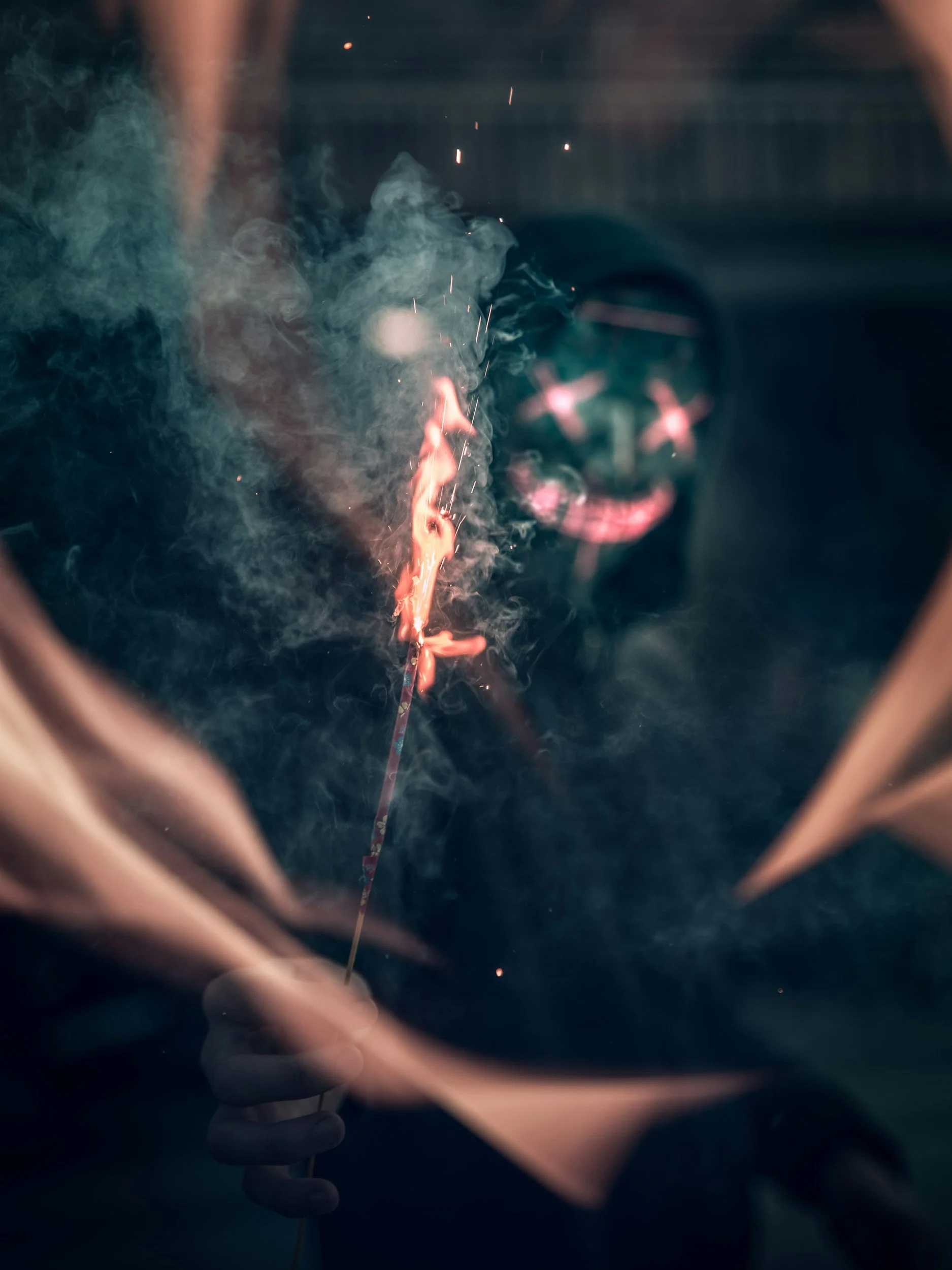 Close-up of a hand holding a lit sparkler in front of a dark background, with sparks and smoke visible.