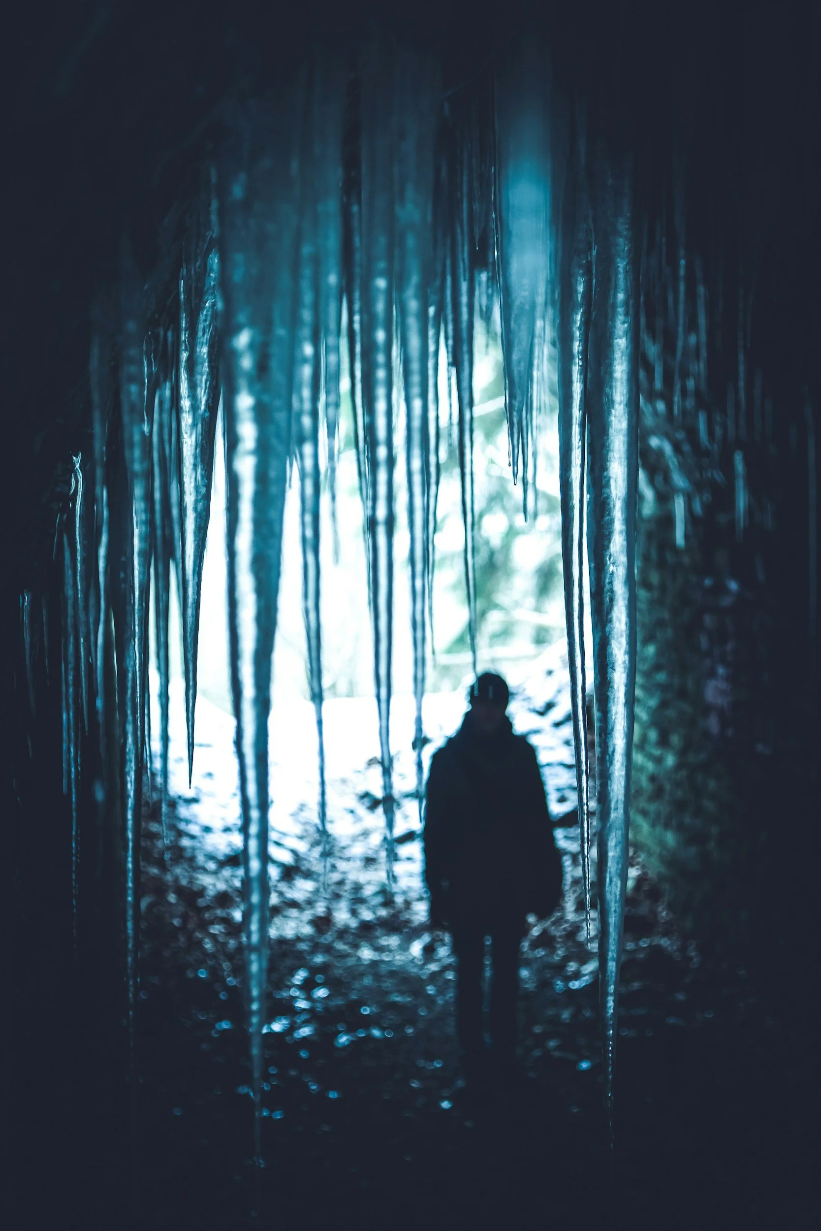 A person sitting inside a dark cave hanging from a ceiling of blue icy stalactites with sunlight streaming in from outside, illuminating the scene.