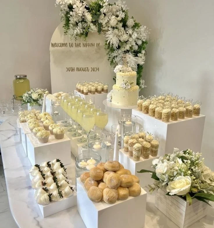 Elegant wedding dessert table with a white theme, decorated with white flowers, a small two-tiered cake, cupcakes, cream puffs, and drinks. A floral arrangement and candles add to the decor, with a backdrop that reads "Welcome to the Nikah of" and the date "20th August 2024."