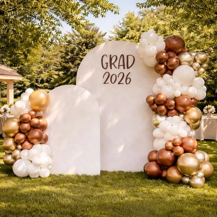 Graduation celebration backdrop with white and bronze balloons surrounding large white circles that read "Grad 2026" on a grassy outdoor setting with trees in the background.