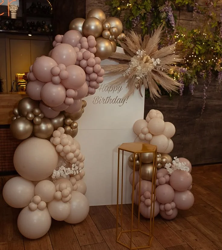 Balloon arrangement with pink, beige, and gold balloons, a white sign with 'Happy Birthday!', dried floral decor, and a gold-colored stand in front, set against a wooden wall and floor for a birthday celebration.