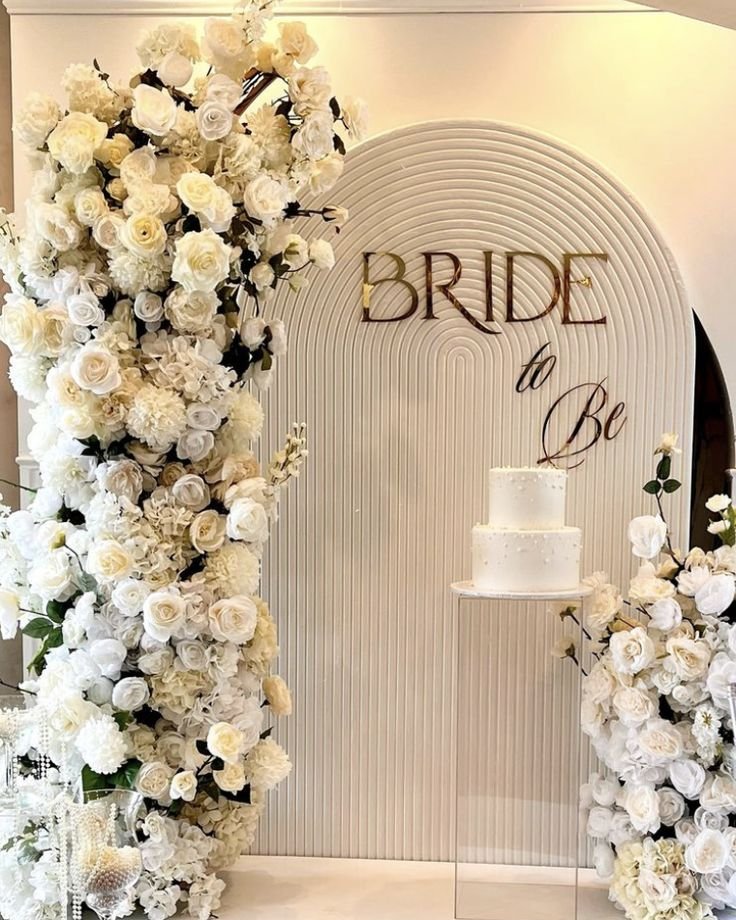 Elegant bridal shower setup with white flowers and a two-tiered wedding cake, featuring a backdrop with a sign reading 'Bride to Be'.