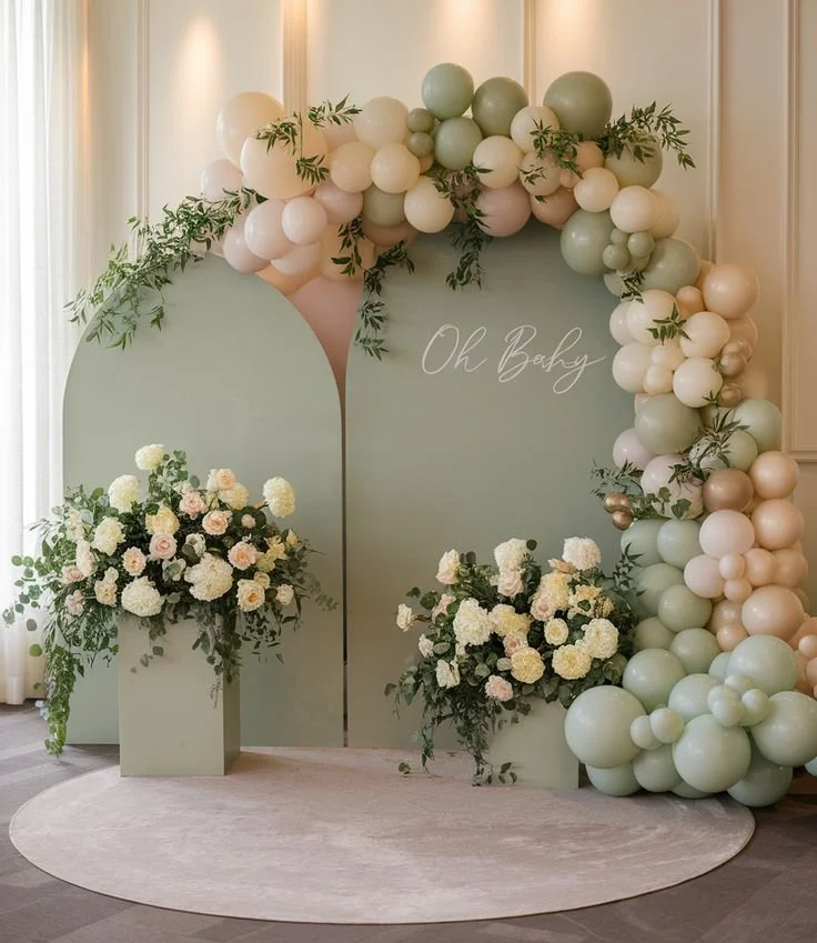 Decorative backdrop with pastel green, beige, and white balloons arranged in an arch, accompanied by floral arrangements of white and blush roses with greenery, on light-colored walls and a round beige rug.