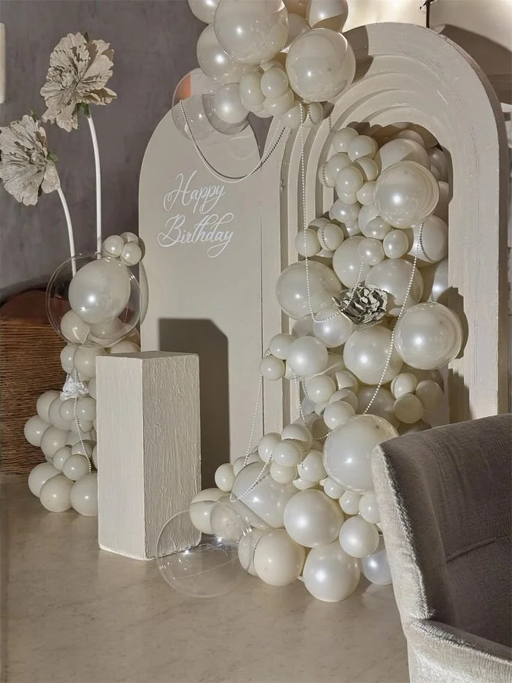 Decorative birthday setup with white balloons, pearl strands, a "Happy Birthday" sign, and floral accents in soft, neutral tones.