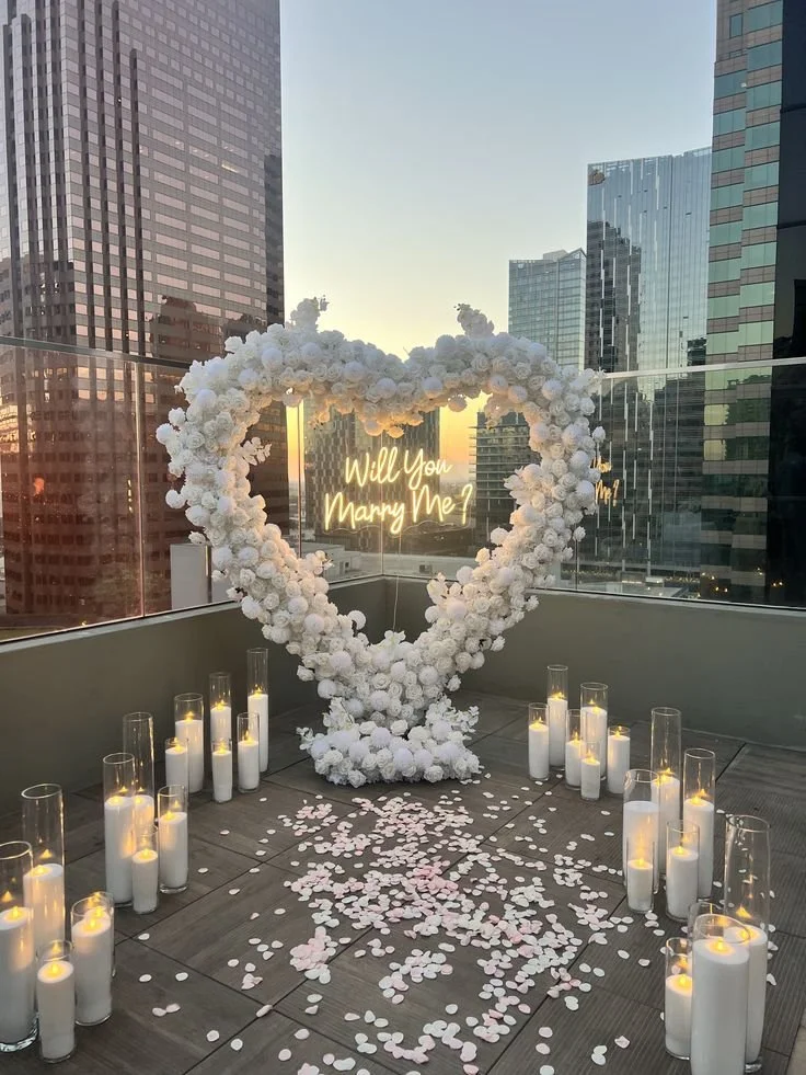 A rooftop setup with a heart-shaped floral arch made of white flowers, surrounded by floating candles in glass holders. Inside the arch, there is a neon sign that says "Will You Marry Me?" in yellow. Petals are scattered on the floor, and the backdrop features a city skyline with tall buildings in the evening sky.