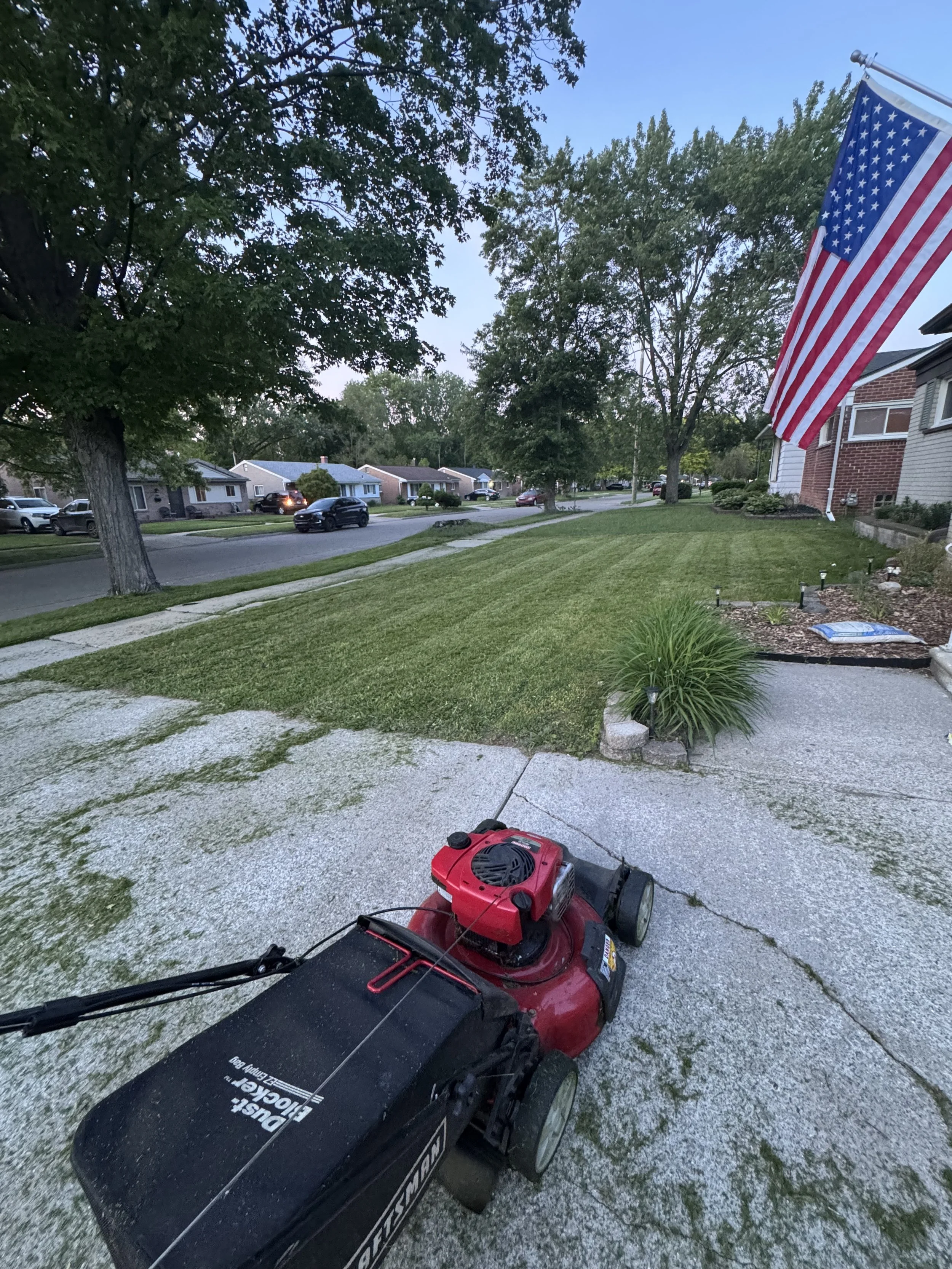 A suburban front yard with green grass, trees, a sidewalk, and a brick house. An American flag is hanging next to the house. A red lawnmower is on the driveway in the foreground.