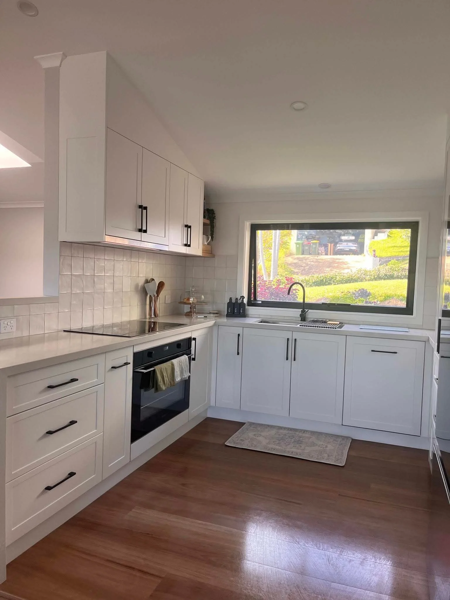Modern kitchen with white cabinets, black handles, a window above the sink letting in natural light, and a wooden floor.