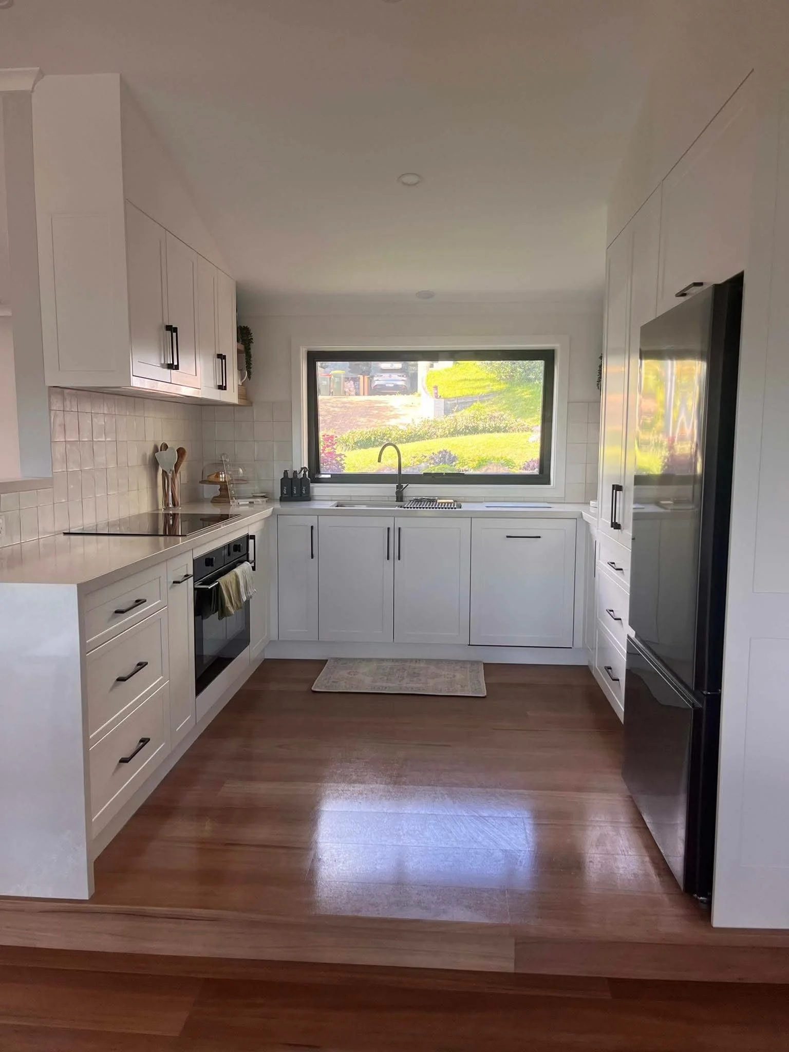 Modern white kitchen with large window, wooden floor, built-in appliances, and minimal decor.