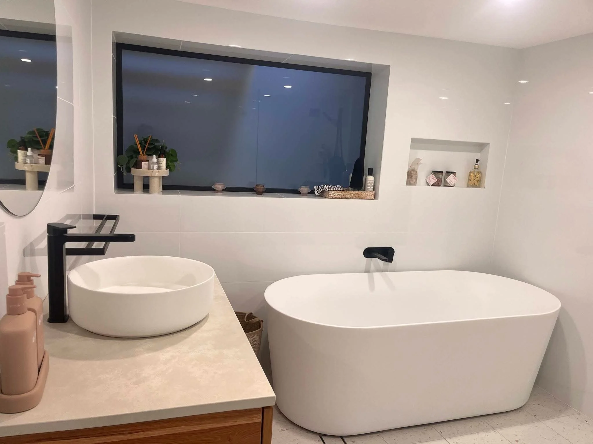 Modern bathroom with a freestanding bathtub, a round vessel sink on a beige countertop, a black faucet, a mirror, and a window with decorative and skincare items on the window ledge and shelf.