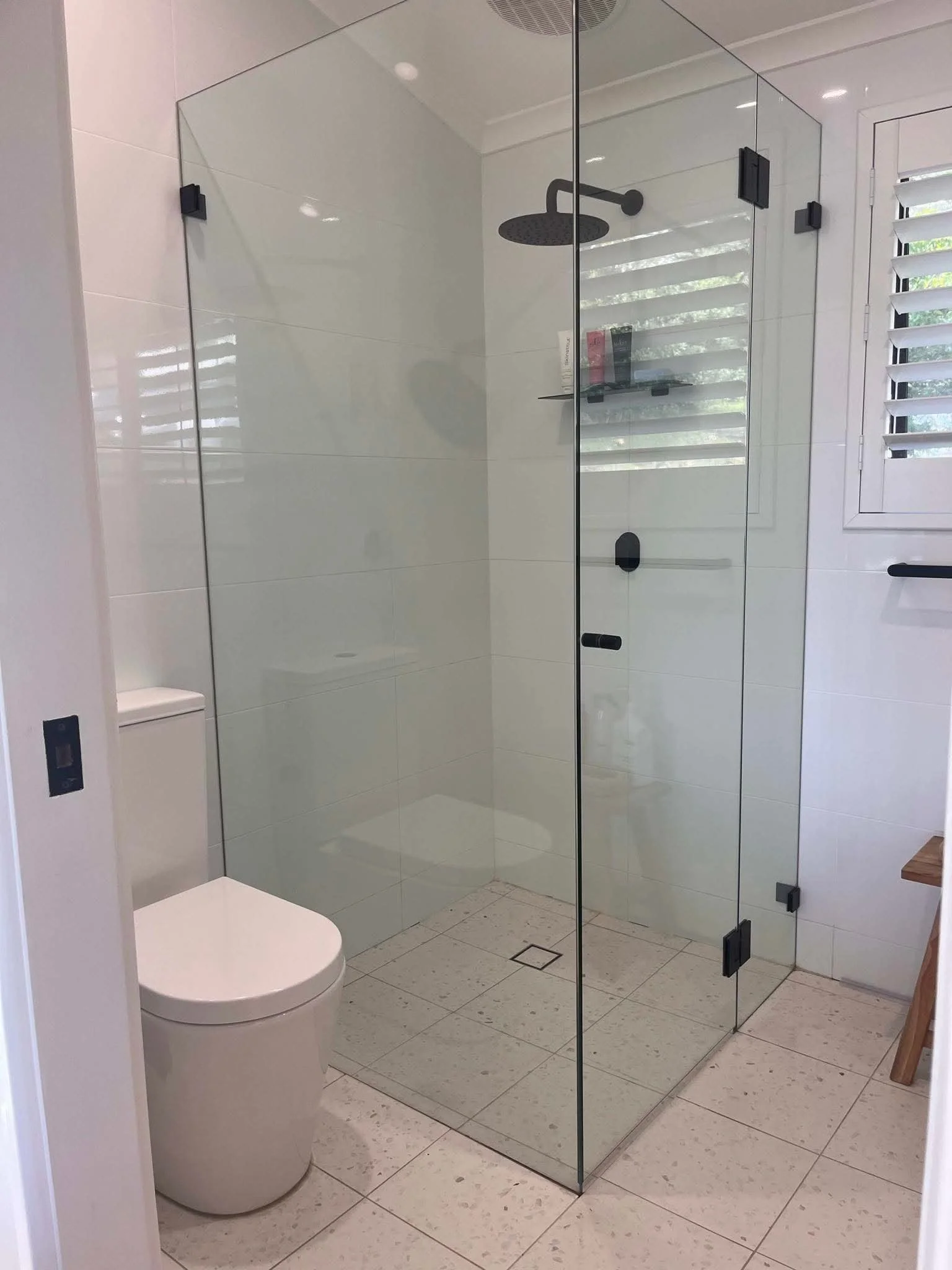 A modern bathroom with a glass shower enclosure, wall-mounted black showerhead, white toilet, tiled walls, and a window with white shutters.
